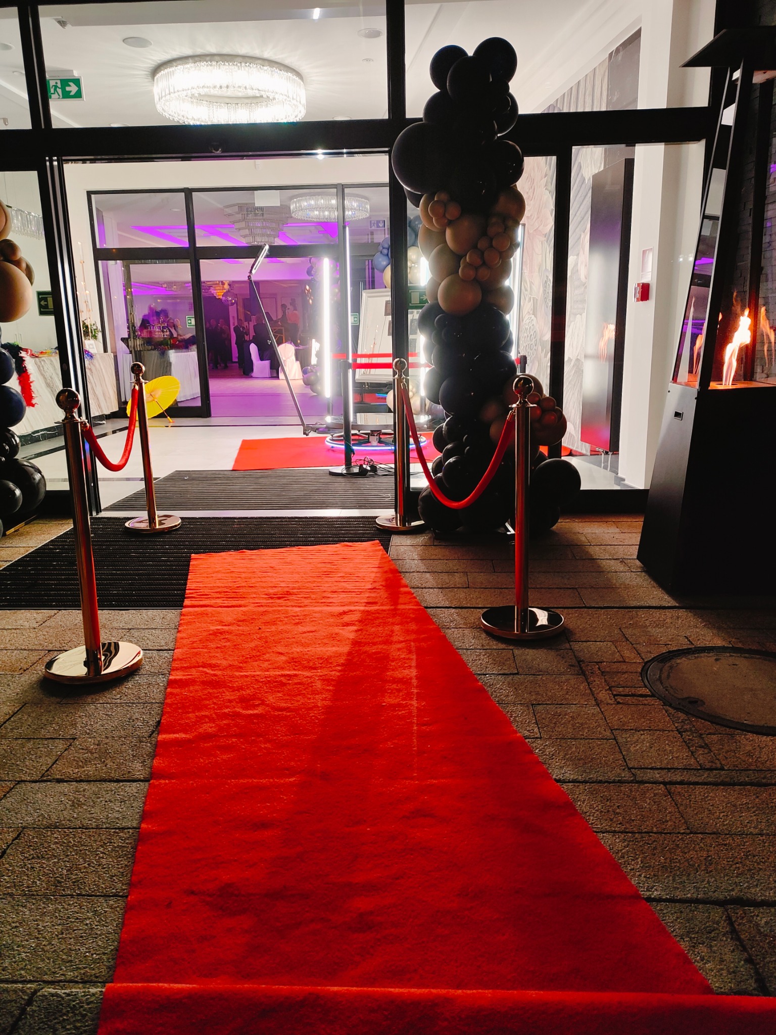 Czerwony dywan prowadzący do wejścia na imprezę, z dekoracją z czarnych i złotych balonów oraz grzejnikiem tarasowym z płomieniami.