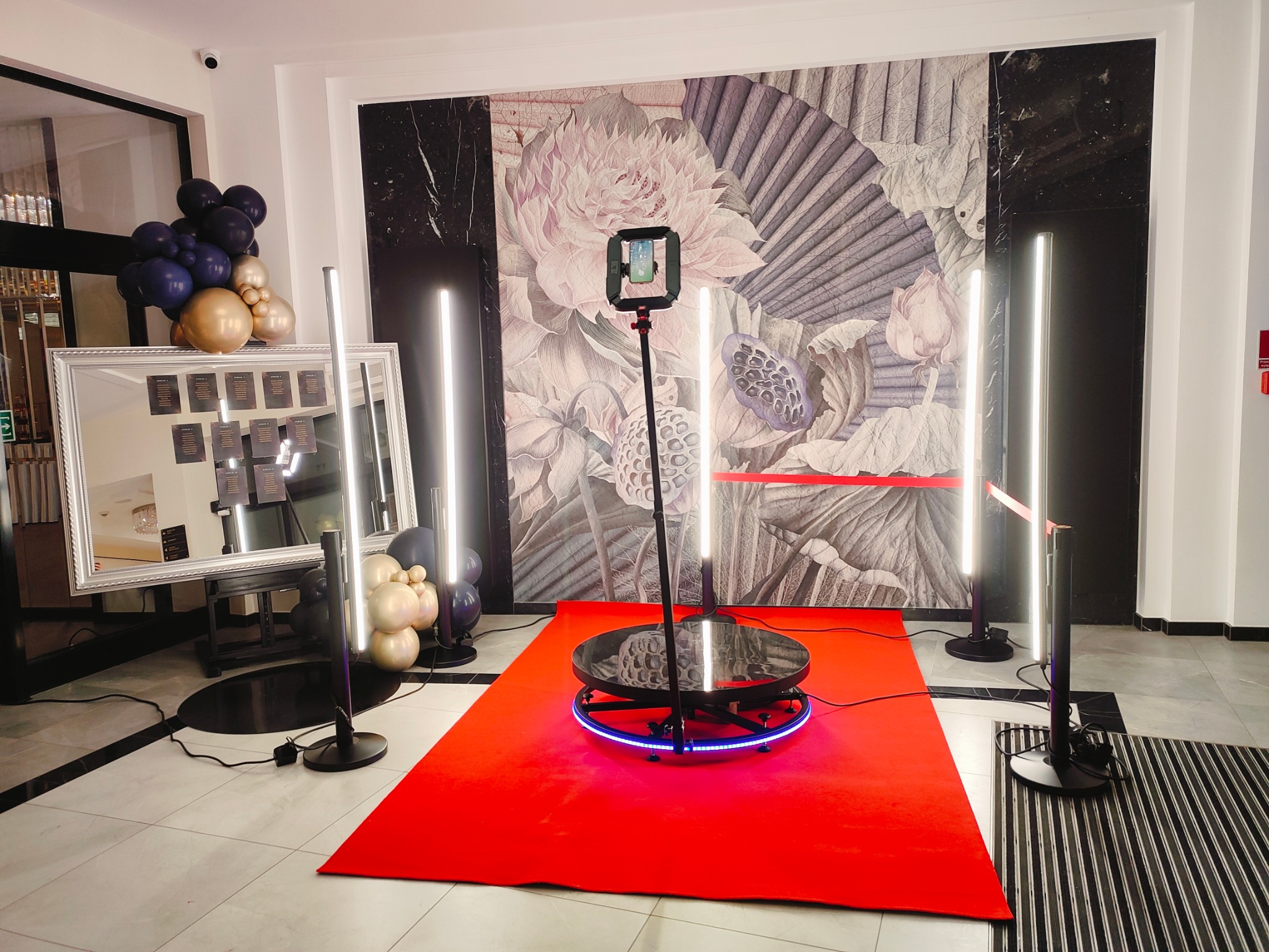 Profesjonalna fotobudka 360 z obrotową platformą LED na czerwonym dywanie, oświetlona lampami LED, tło z motywem kwiatowym i dekoracjami balonowymi.