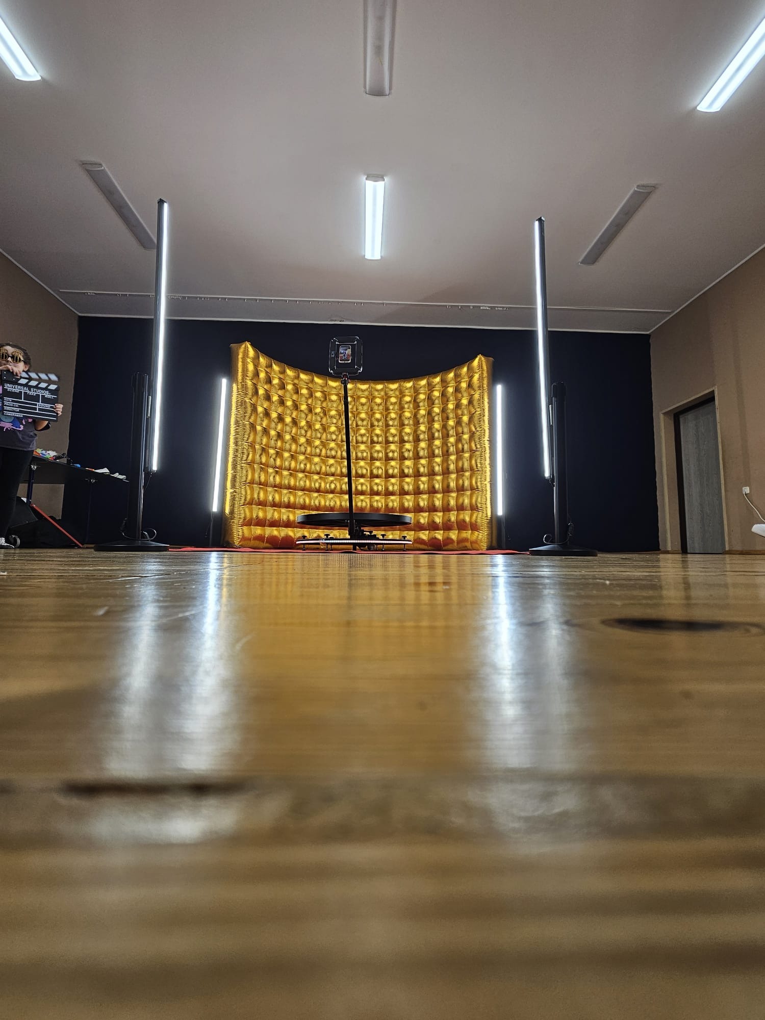 Złota, dmuchana ścianka fotobudki ze smartfonem na statywie i oświetleniem LED, z klapsem filmowym w tle, widziana z perspektywy podłogi.