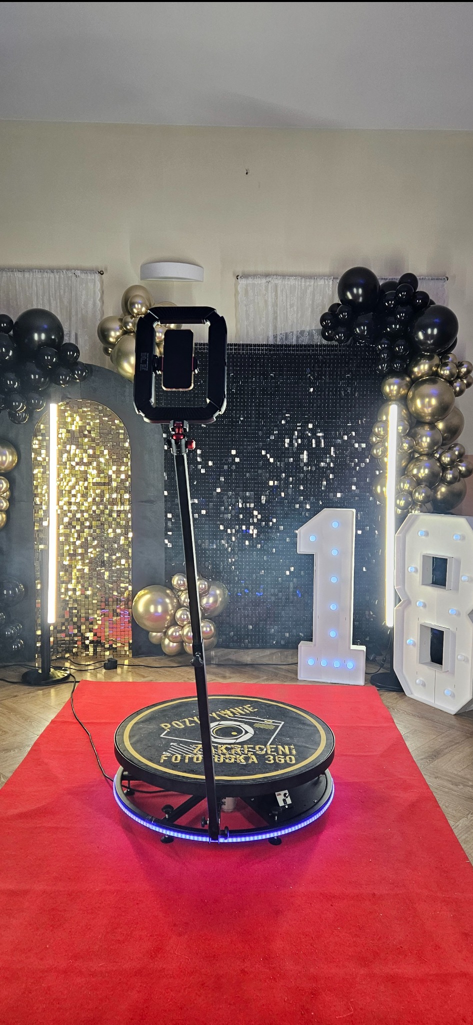 Okrągła fotobudka 360 na czerwonym dywanie, z dekoracjami balonowymi i podświetlanymi cyframi '1' i '8' w tle, tworząca imprezową atmosferę.