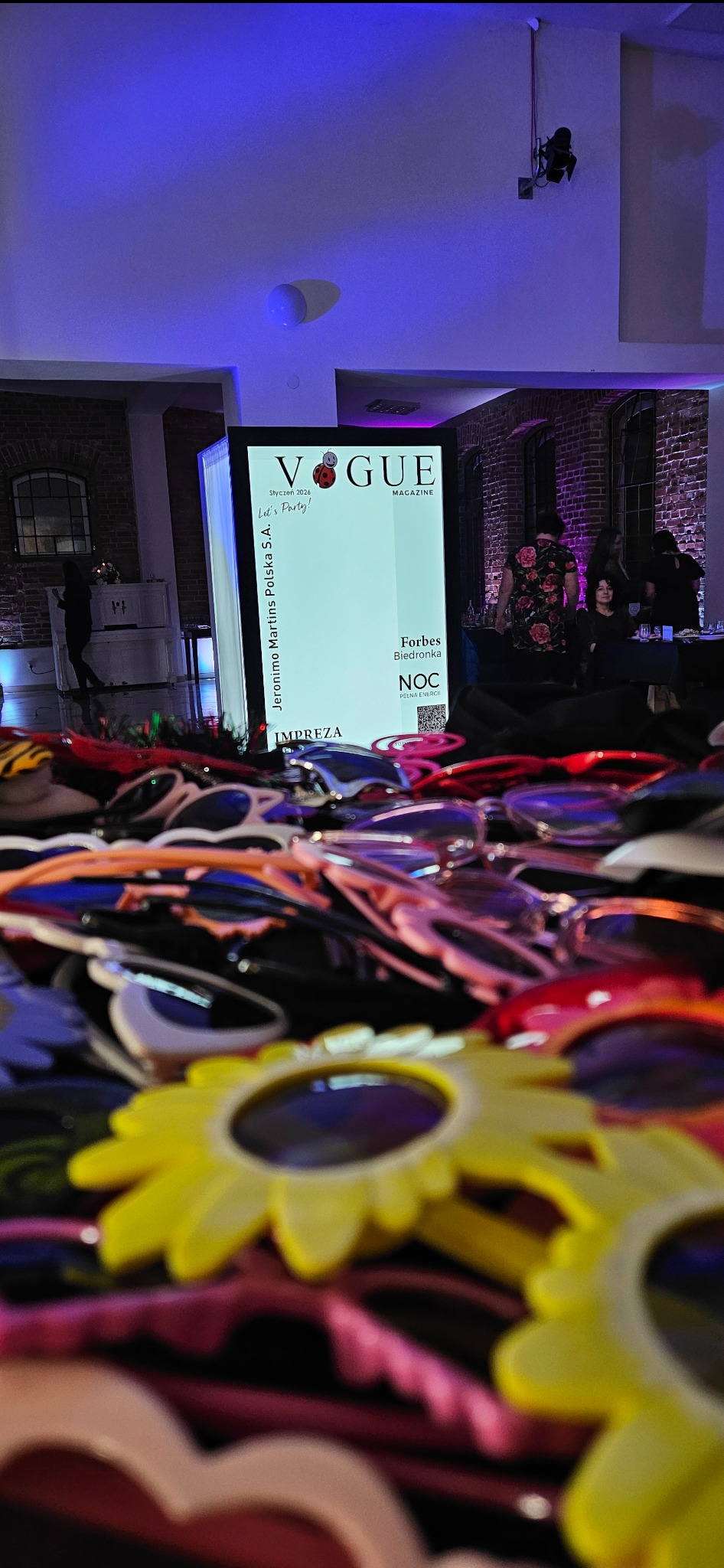 Atrakcje na imprezie: okulary w kształcie kwiatów na pierwszym planie, w tle fotobudka z logo Vogue Magazine i goście. Ceglana ściana w tle, niebieskie oświetlenie.