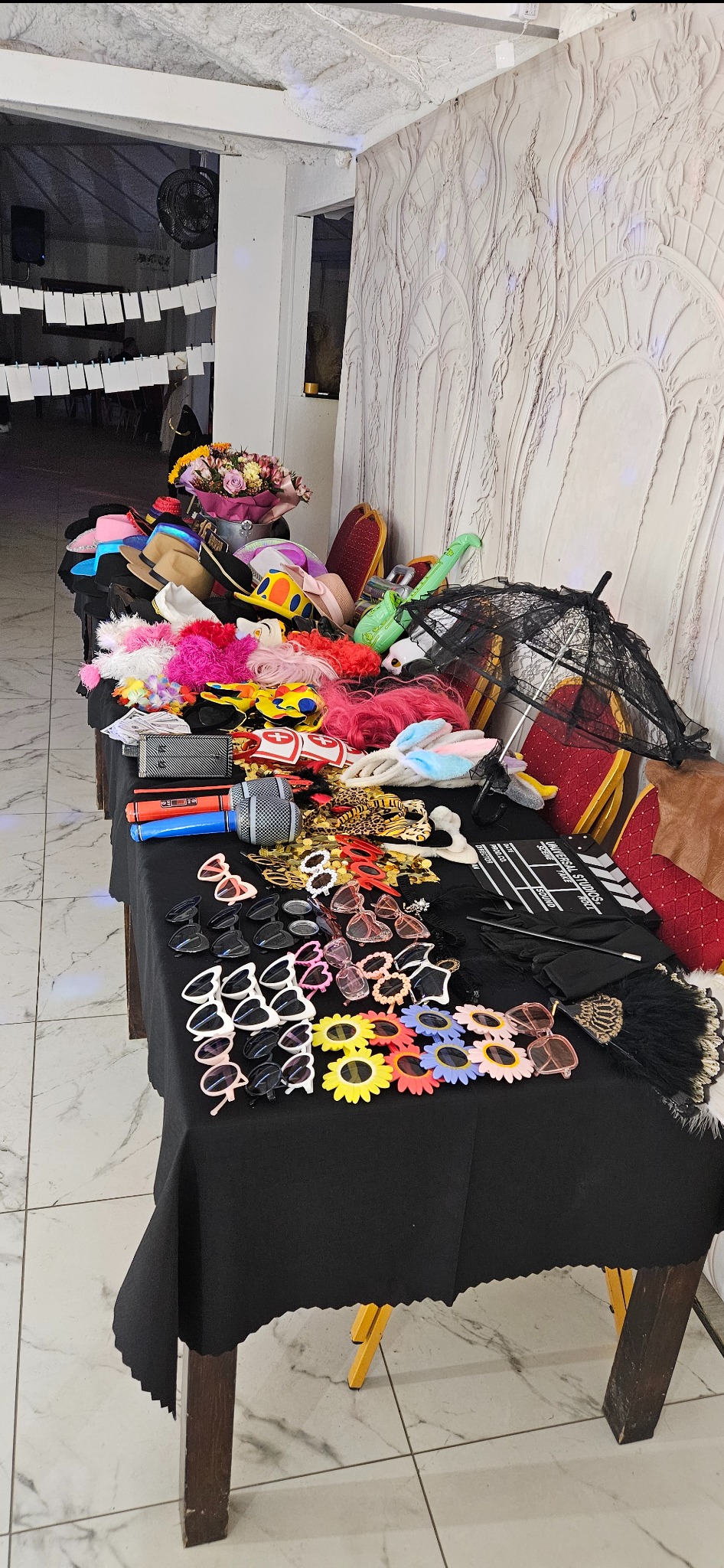 Stół z rekwizytami do sesji zdjęciowej: kapelusze, okulary, peruki, mikrofon, klaps filmowy. Dekoracyjne tło i kwiaty. Różnorodność akcesoriów na czarnym obrusie.