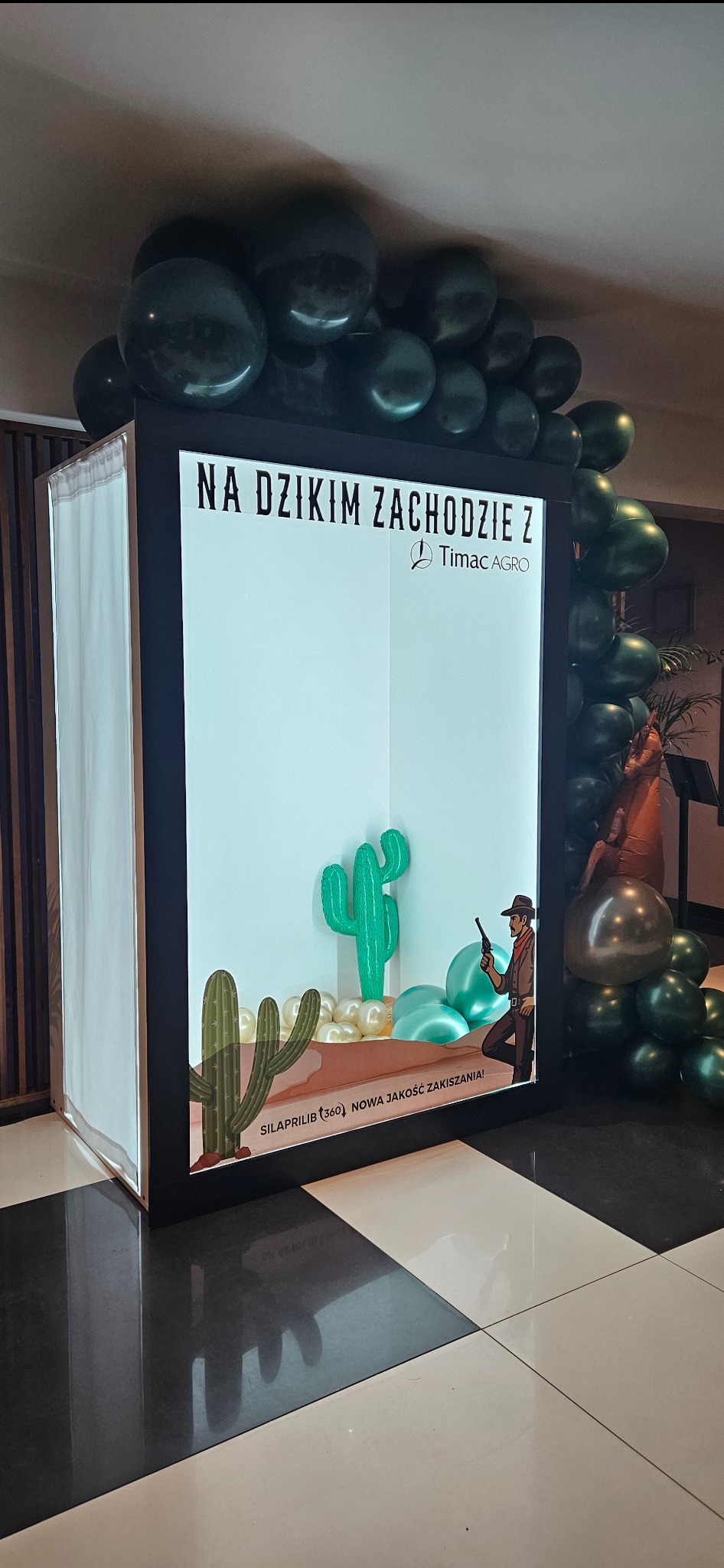 Tematyczna ścianka do zdjęć 'Na Dzikim Zachodzie' z balonami, kaktusami i postacią kowboja, ustawiona na czarno-białej podłodze.
