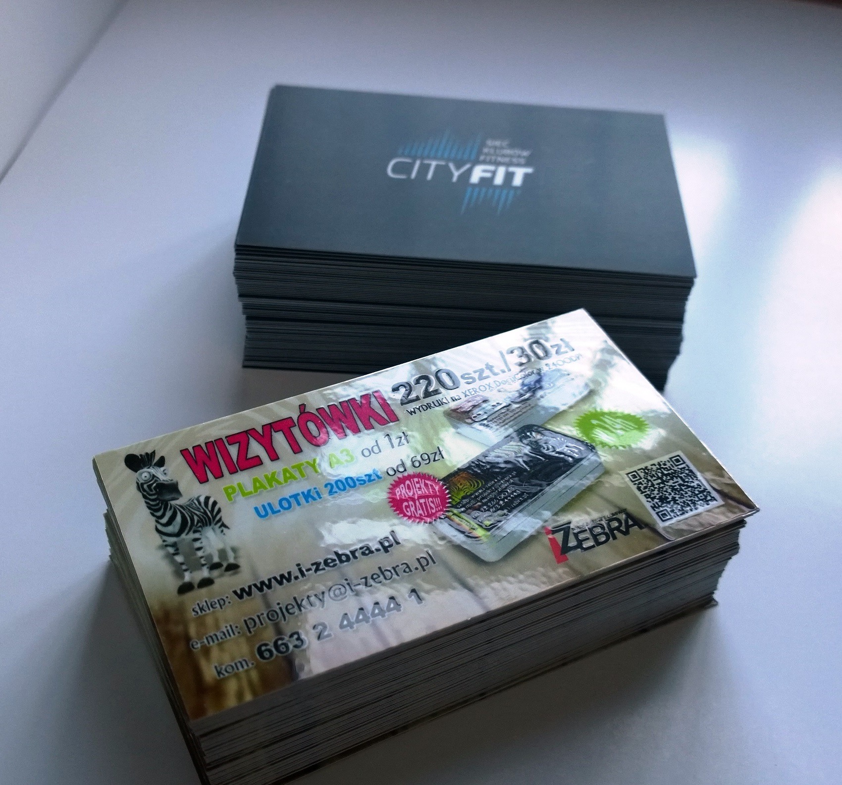 Dwie stosy wydrukowanych materiałów reklamowych. Na wierzchu stos z informacjami o ofercie firmy i-zebra.pl, z hasłem 'Wizytówki 220 szt. / 30 zł', obok drugi stos z logo CITYFIT.
