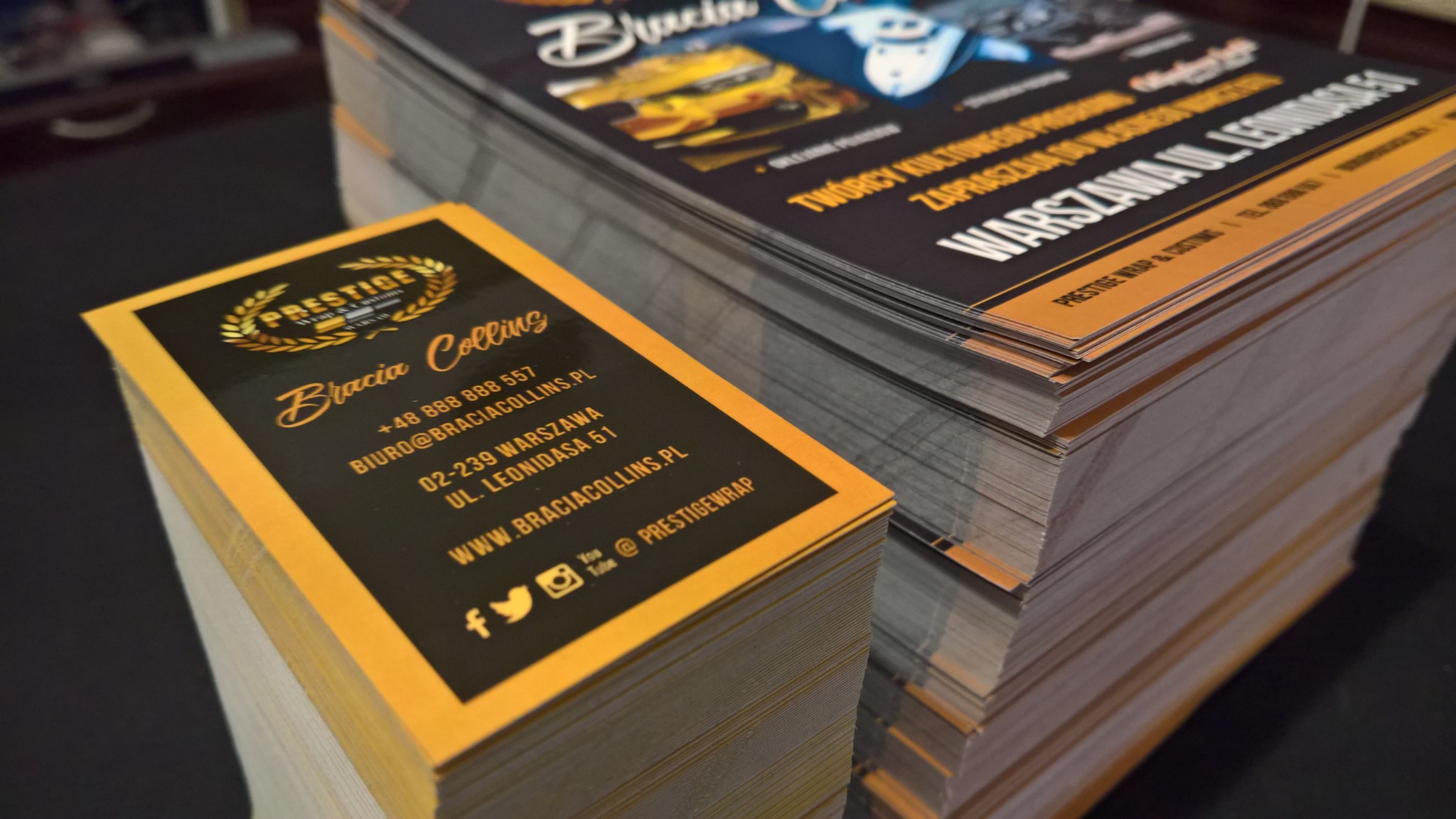 Stosy świeżo wydrukowanych materiałów reklamowych, w tym wizytówki z czarno-złotym motywem i logo firmy, oraz ulotki z grafiką samochodową, leżące na ciemnym blacie.