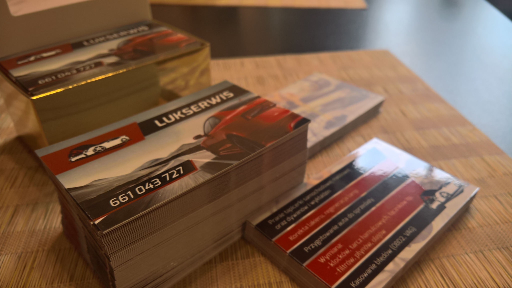 Stosy błyszczących wizytówek firmy 'LUKSERWIS' z grafiką czerwonego samochodu sportowego i danymi kontaktowymi, ułożone na drewnianym blacie.