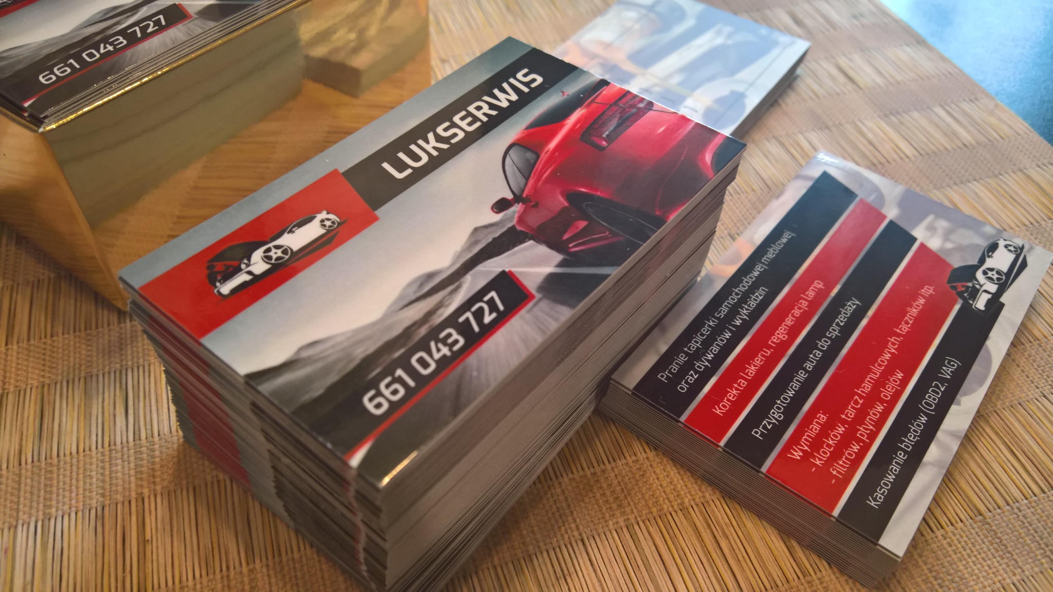 Stosy wizytówek firmy Lukserwis z grafiką czerwonego samochodu i danymi kontaktowymi, leżące na plecionym podkładzie.