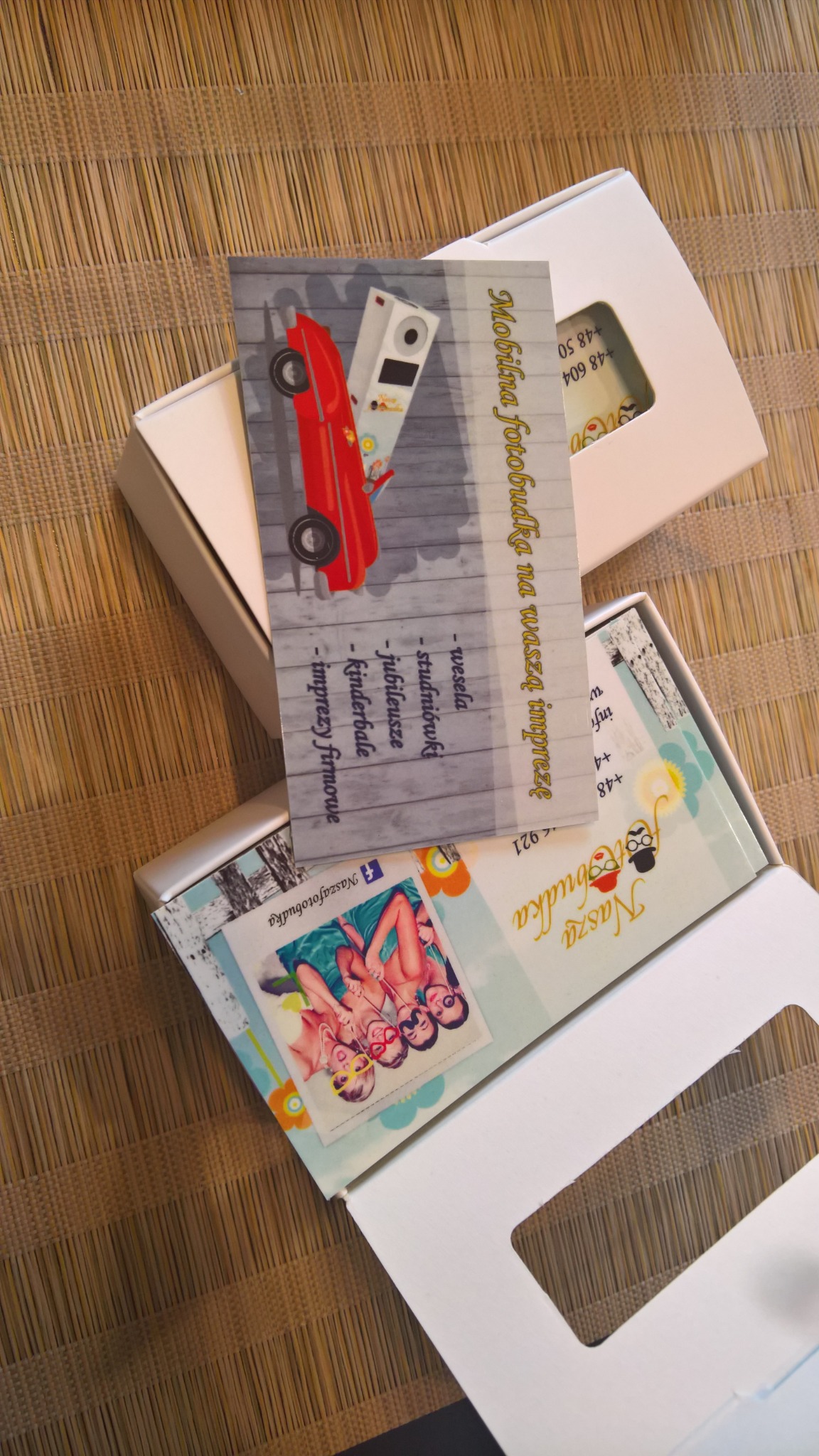 Dwa pudełka z wizytówkami fotobudki, jedna z grafiką czerwonego samochodu i aparatu, druga z grupą osób w strojach kąpielowych, leżące na bambusowej macie.