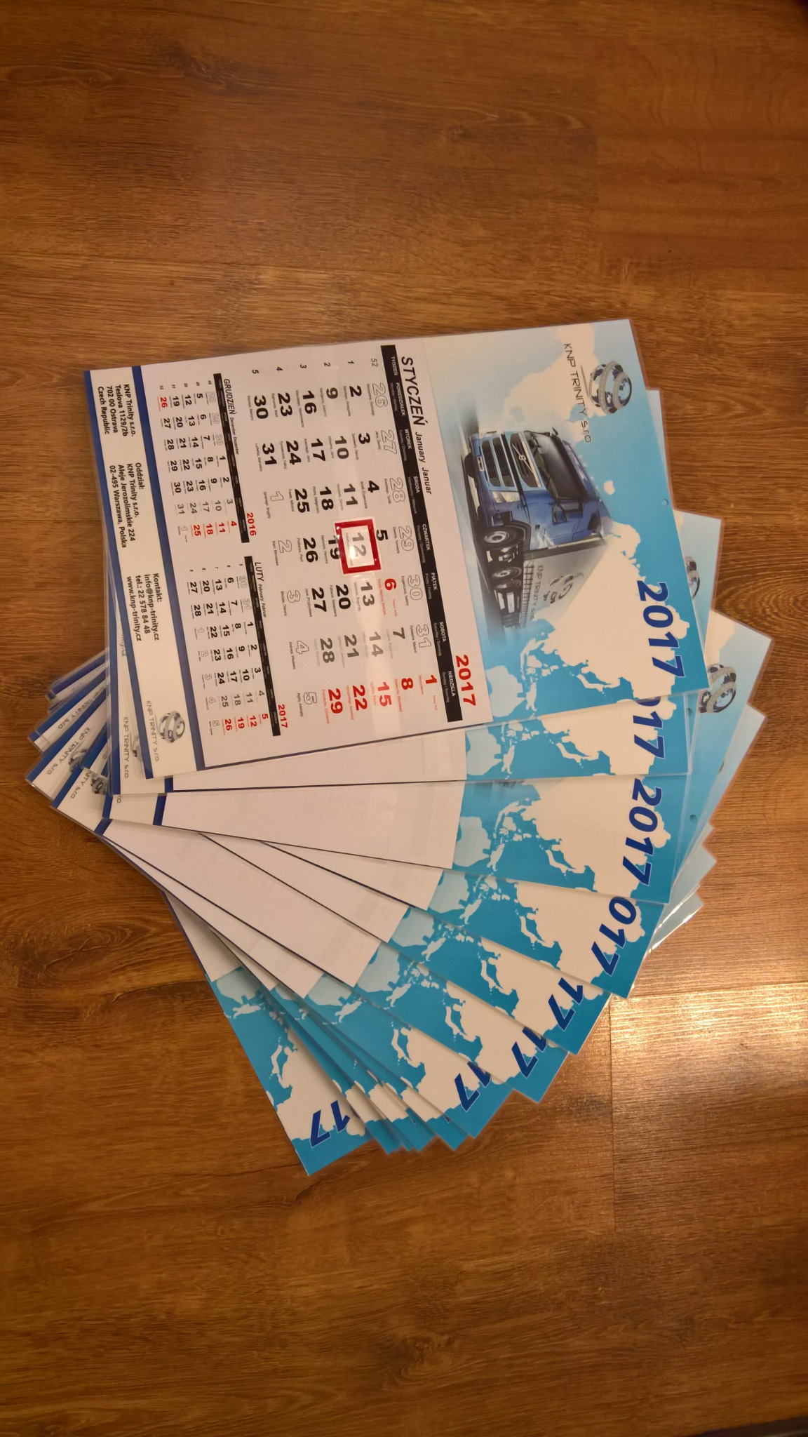Stos firmowych kalendarzy trójdzielnych z grafiką ciężarówki i logo firmy KNP Trinity S.r.o., widoczne daty z zaznaczonym dniem.