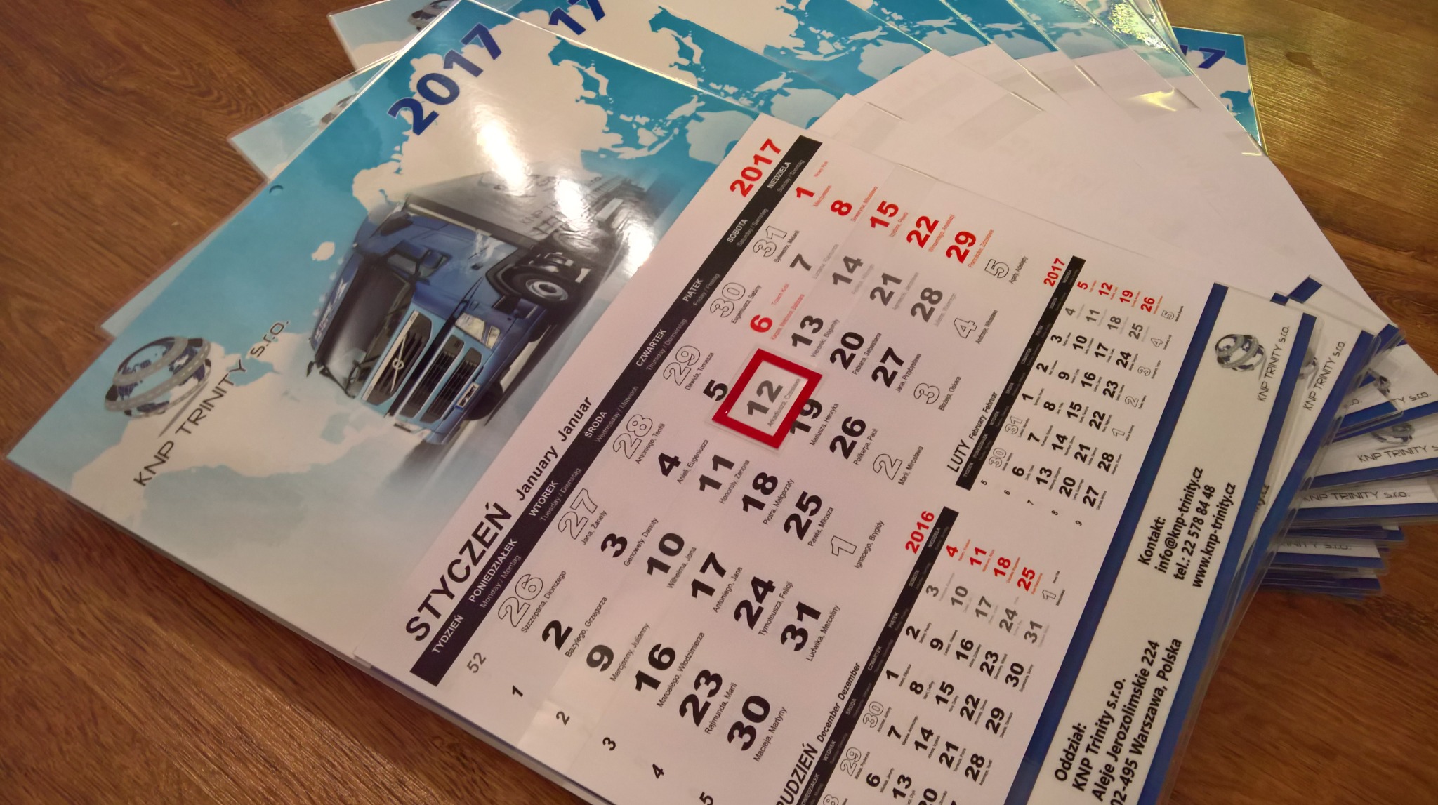 Stos kalendarzy ściennych z motywem transportowym i logo firmy KNP Trinity s.r.o., widoczne strony z miesiącem styczeń 2017.
