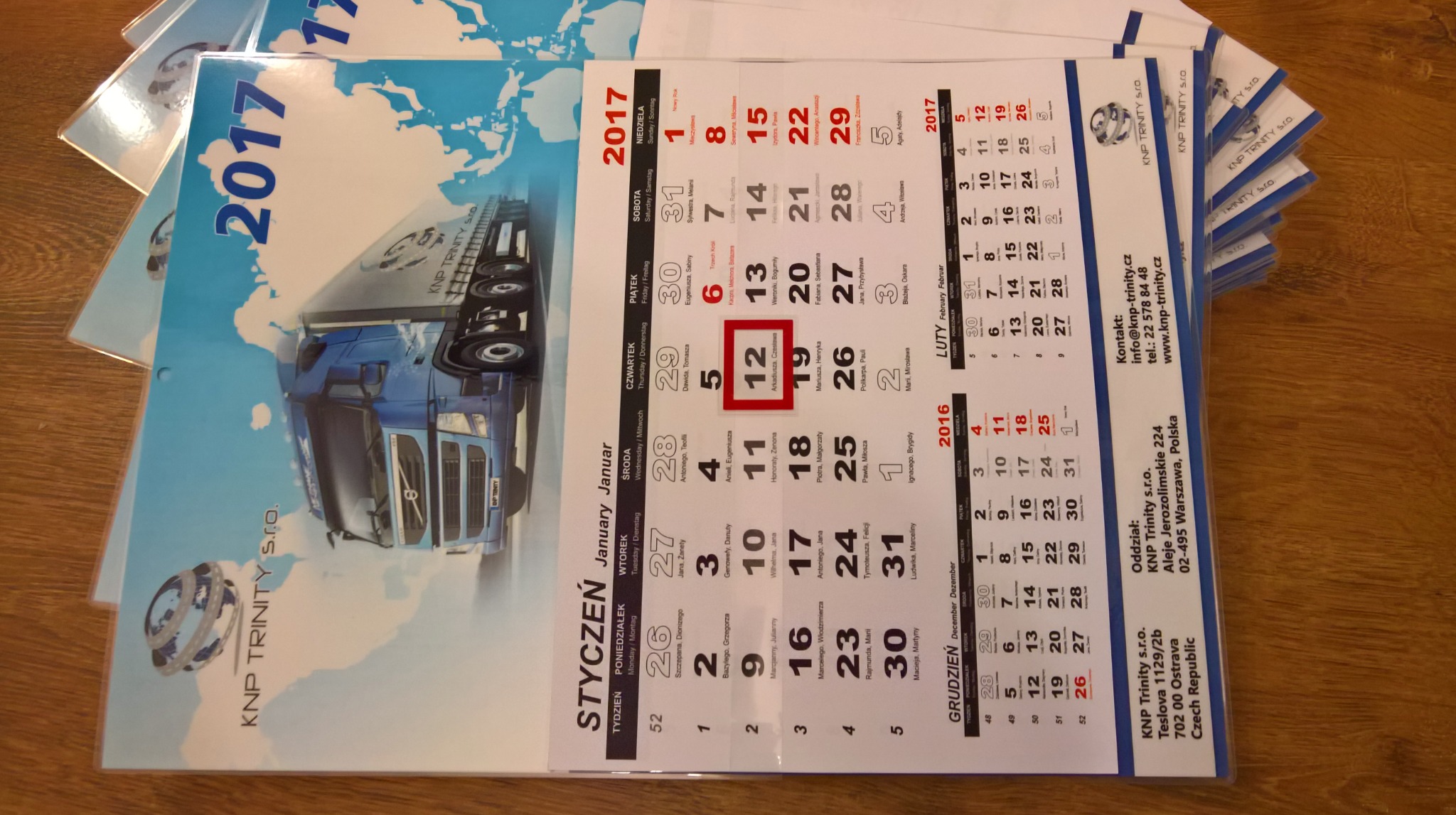 Stos firmowych kalendarzy ściennych z ilustracją niebieskiej ciężarówki na tle mapy świata, rok 2017, widoczne daty w języku polskim.