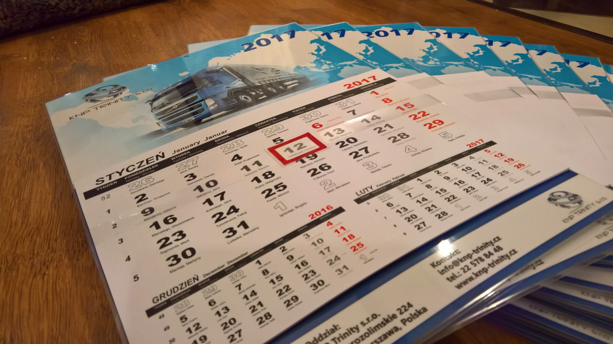 Stos kalendarzy firmowych na rok 2017 z widocznym motywem graficznym ciężarówki na tle błękitnego nieba, leżących na drewnianej powierzchni.