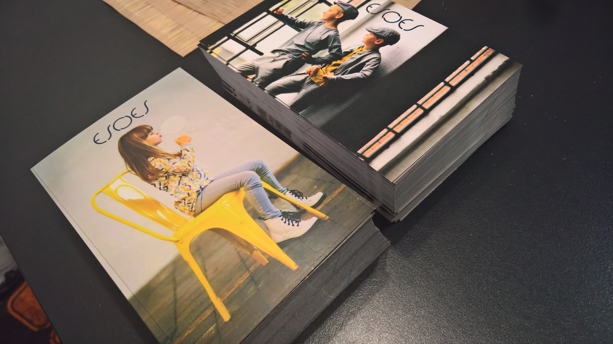 Stosy świeżo wydrukowanych katalogów z różnymi okładkami na czarnym tle, widoczne detale graficzne i tekst na okładkach.