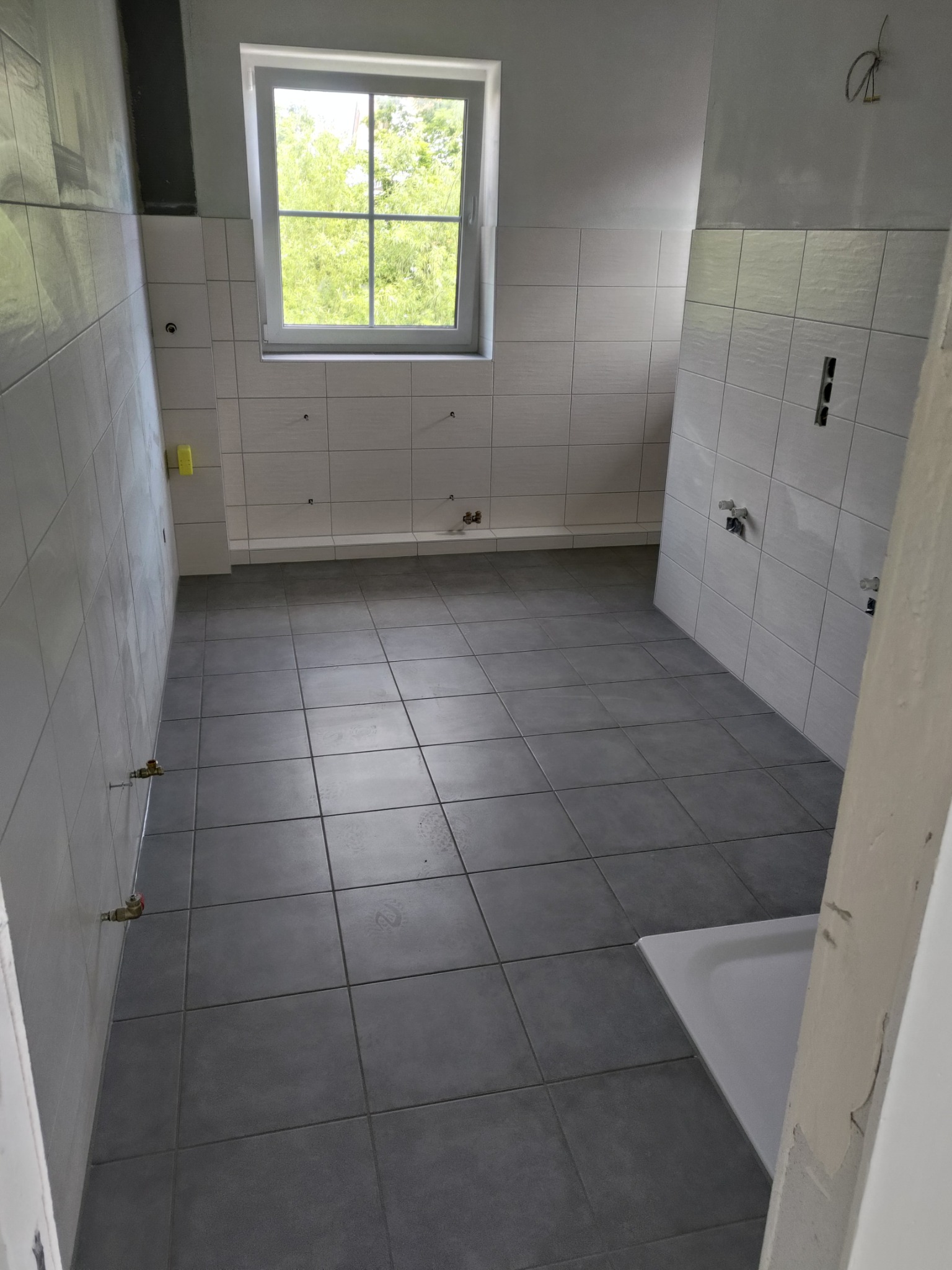 Wnętrze łazienki w trakcie prac wykończeniowych: szare płytki podłogowe, białe płytki ścienne do połowy wysokości, okno z widokiem na zieleń, przygotowane punkty instalacyjne.
