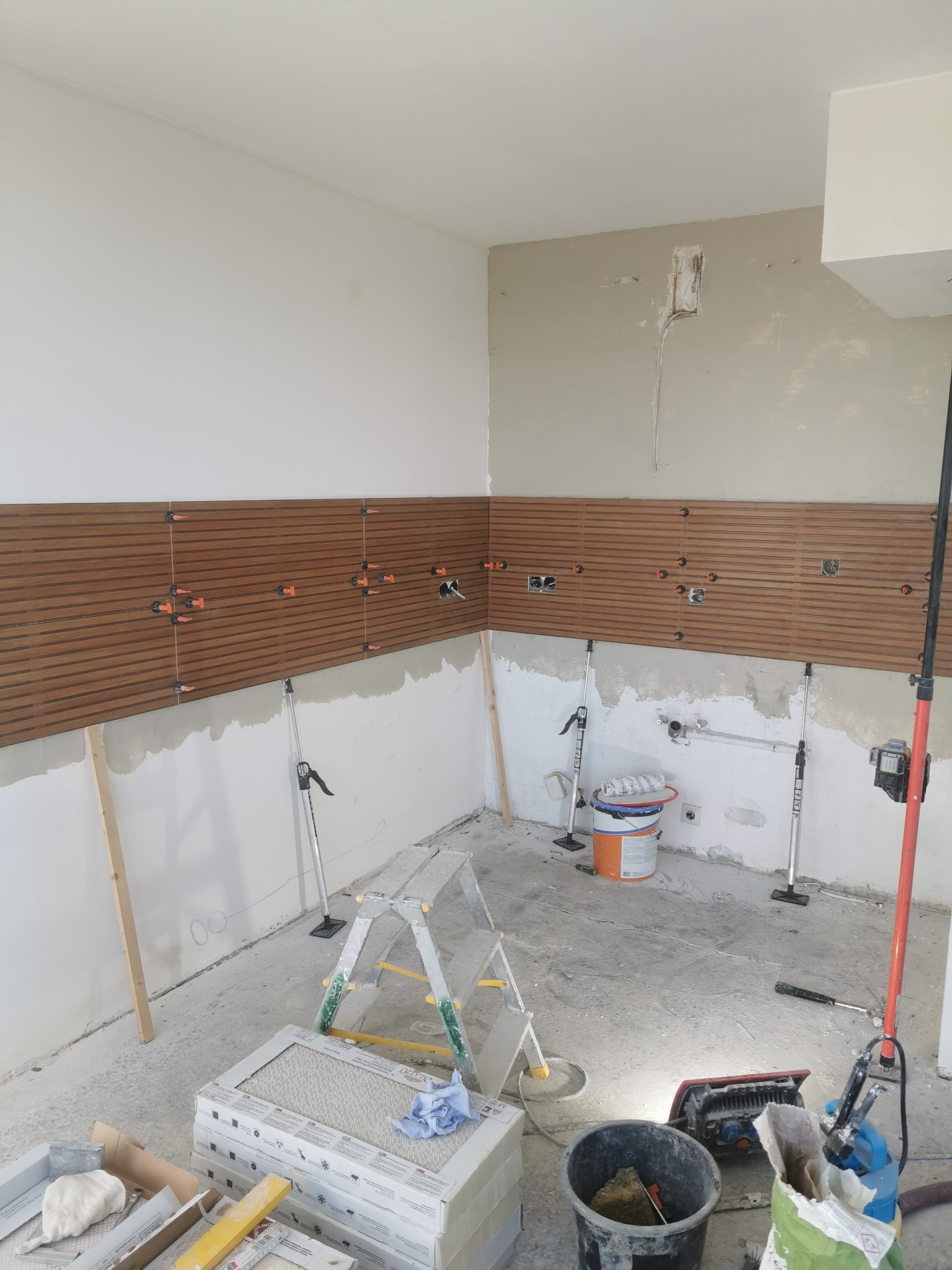 Montaż paneli ściennych imitujących drewno w kuchni, widoczne poziomice laserowe, krzyżyki dystansowe i drabina, ściany przygotowane pod dalsze prace, częściowo położone panele z poziomymi rowkami.