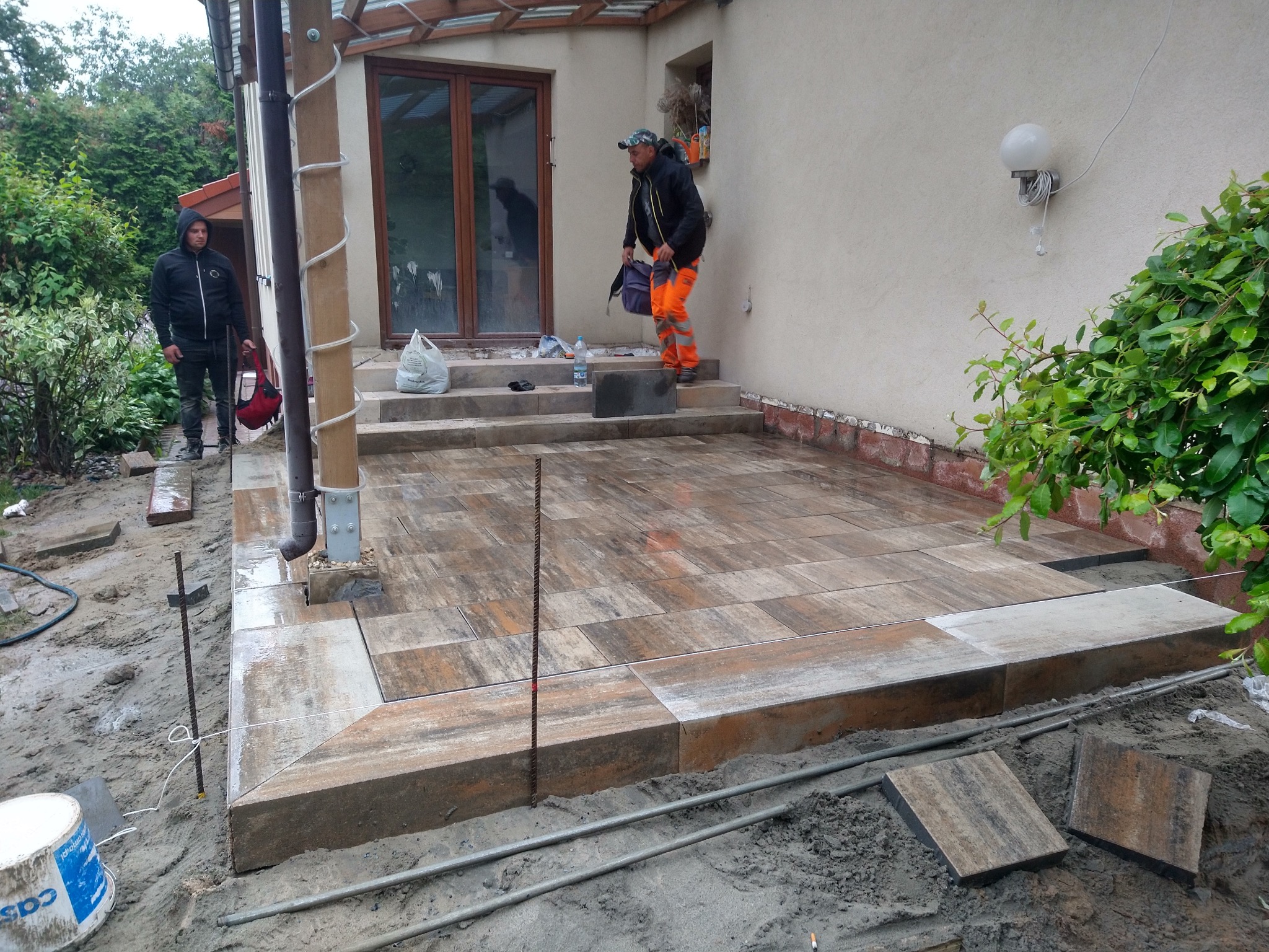 Prace brukarskie w toku: układanie tarasu z brązowo-szarej kostki, widoczni dwaj pracownicy w odzieży roboczej, jeden na schodach, drugi przy krawędzi tarasu, narzędzia i materiały budowlane w tle,...