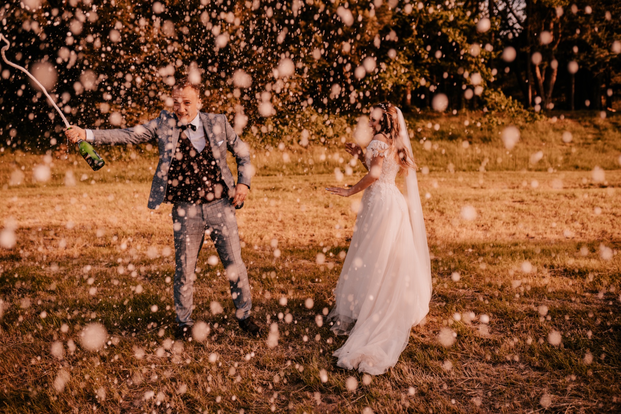 Uśmiechnięty pan młody w szarym garniturze otwiera szampana, zalewając parę młodą fontanną bąbelków na tle zielonego pola i drzew.