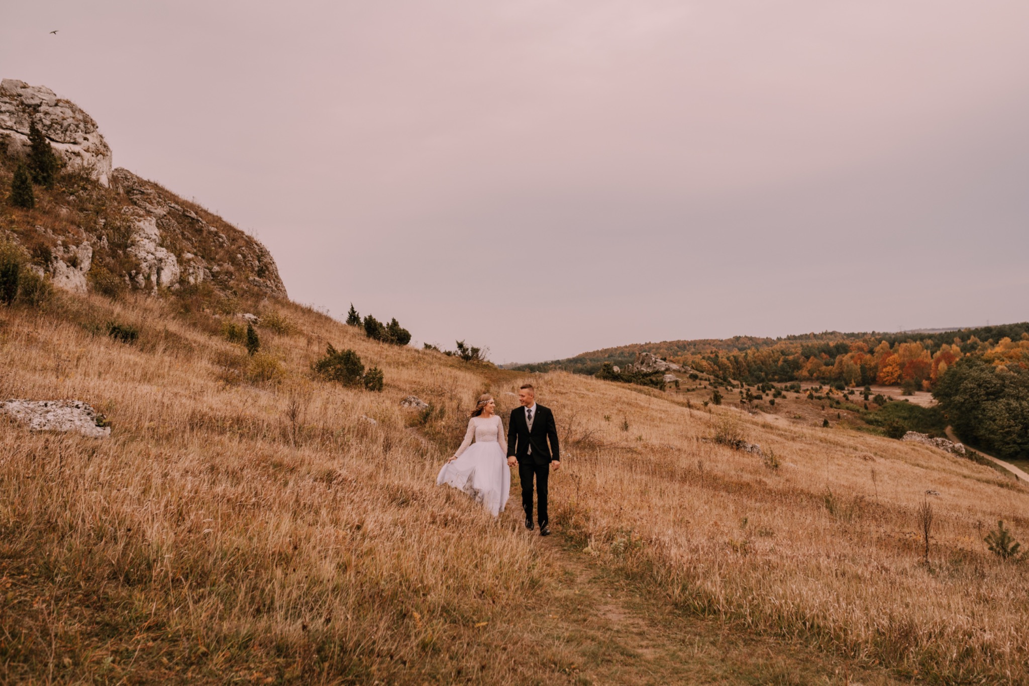 Para młoda, panna młoda w długiej sukni z koronkowymi rękawami i pan młody w czarnym garniturze, spacerują po wzgórzu porośniętym suchą trawą, w tle skały i drzewa w jesiennych kolorach, pochmurne...
