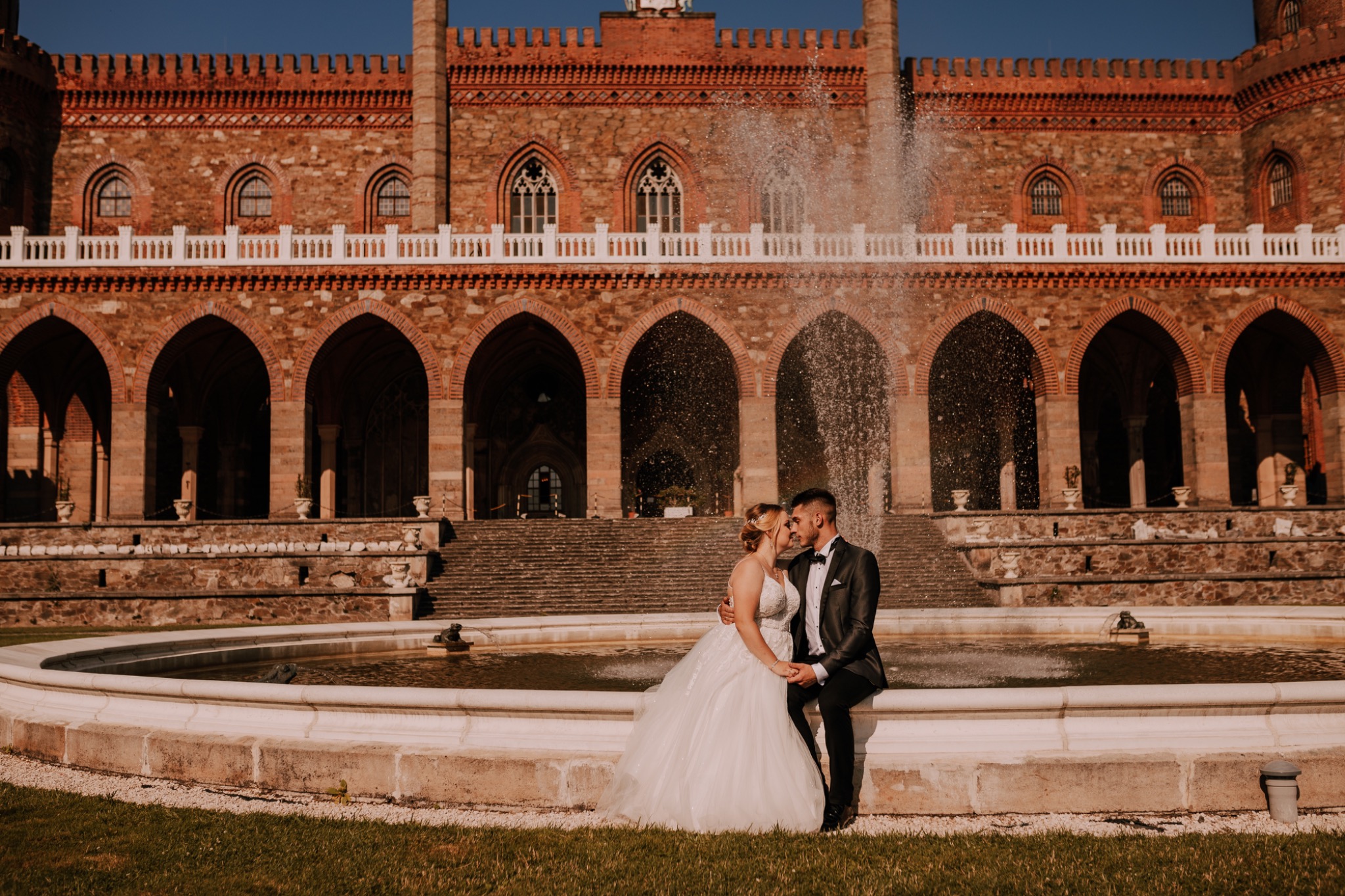 Para młoda siedzi na krawędzi fontanny na tle monumentalnego, ceglanego zamku z arkadami, woda z fontanny tworzy delikatną zasłonę, a para delikatnie się całuje.