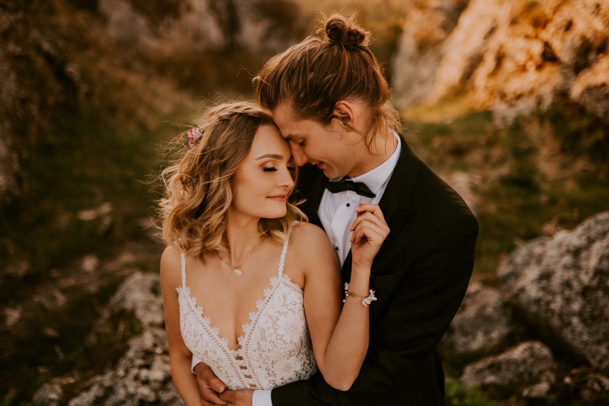 Para młoda w objęciach na tle skalistego krajobrazu: panna młoda w koronkowej sukni z kwiatami we włosach, pan młody w czarnym smokingu z muszką, delikatny uśmiech obojga.