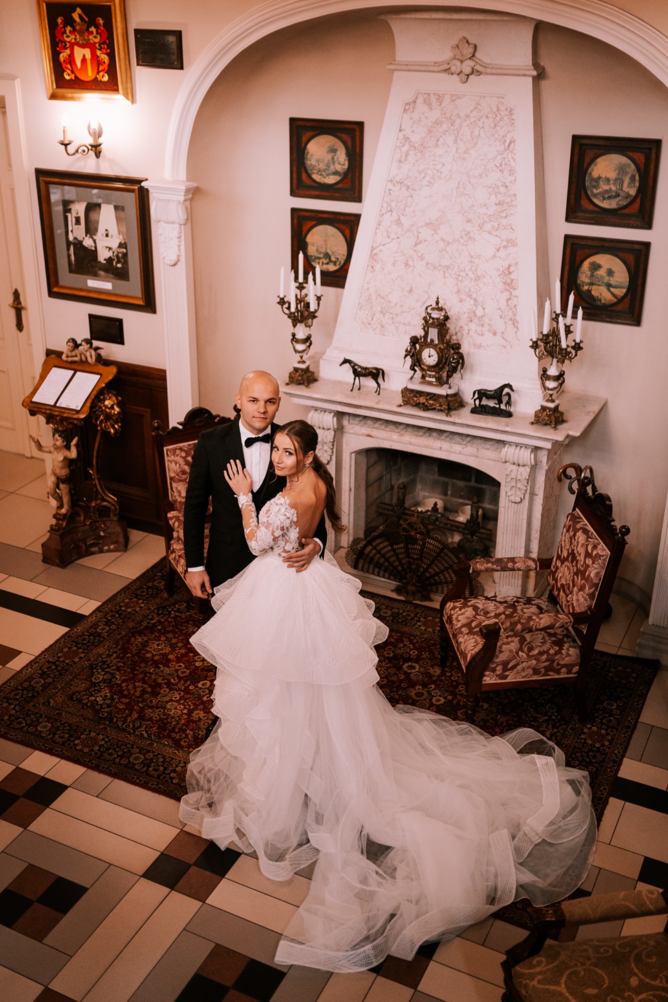 Para młoda w eleganckim wnętrzu z kominkiem, ona w sukni z falbanami i koronkowymi rękawami, on w czarnym smokingu z muchą, widok z góry.