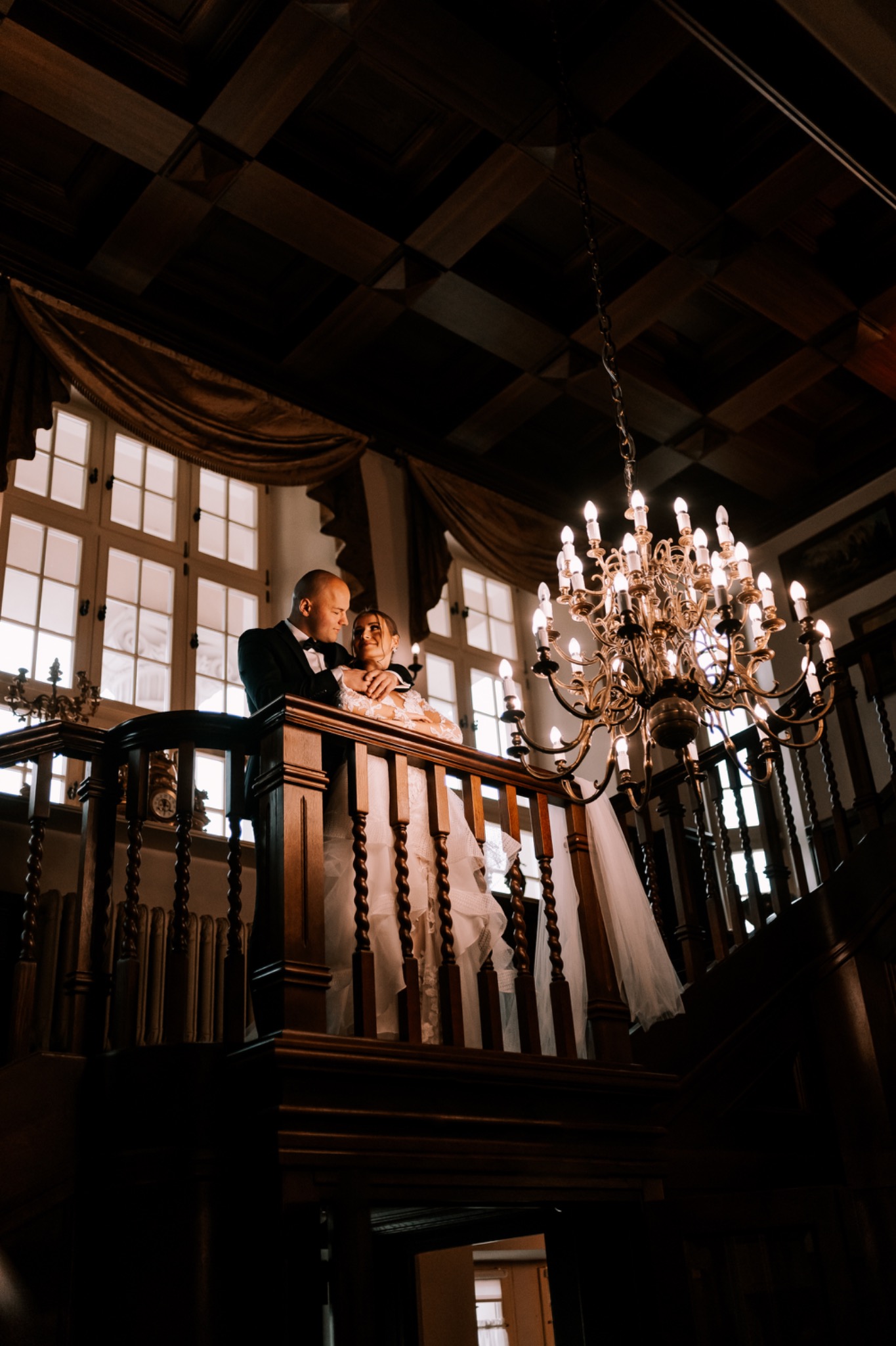 Elegancka para młoda na drewnianej antresoli z balustradą, w tle okna z zasłonami i kryształowy żyrandol