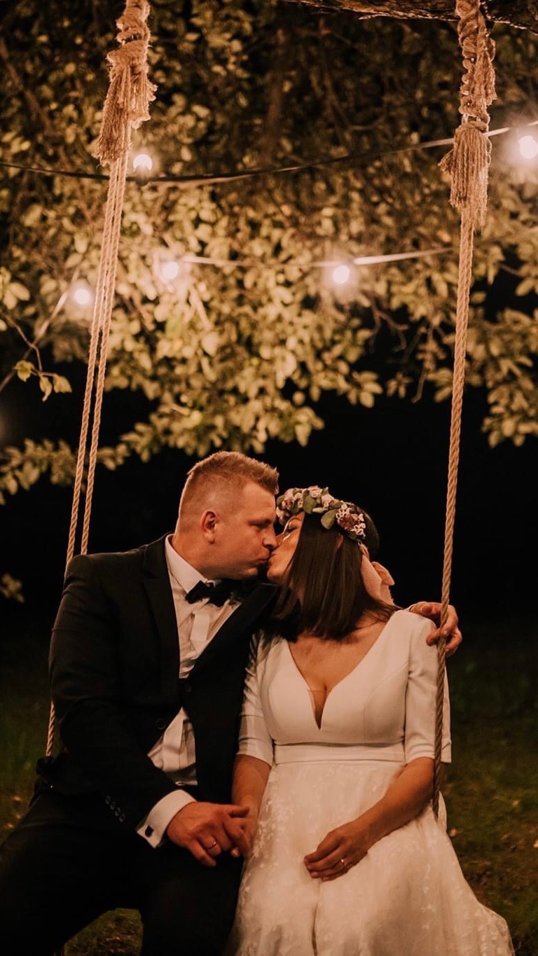 Pocałunek nowożeńców na huśtawce zawieszonej pod drzewem z lampkami, on w czarnym smokingu z muchą, ona w białej sukni z wiankiem na głowie.