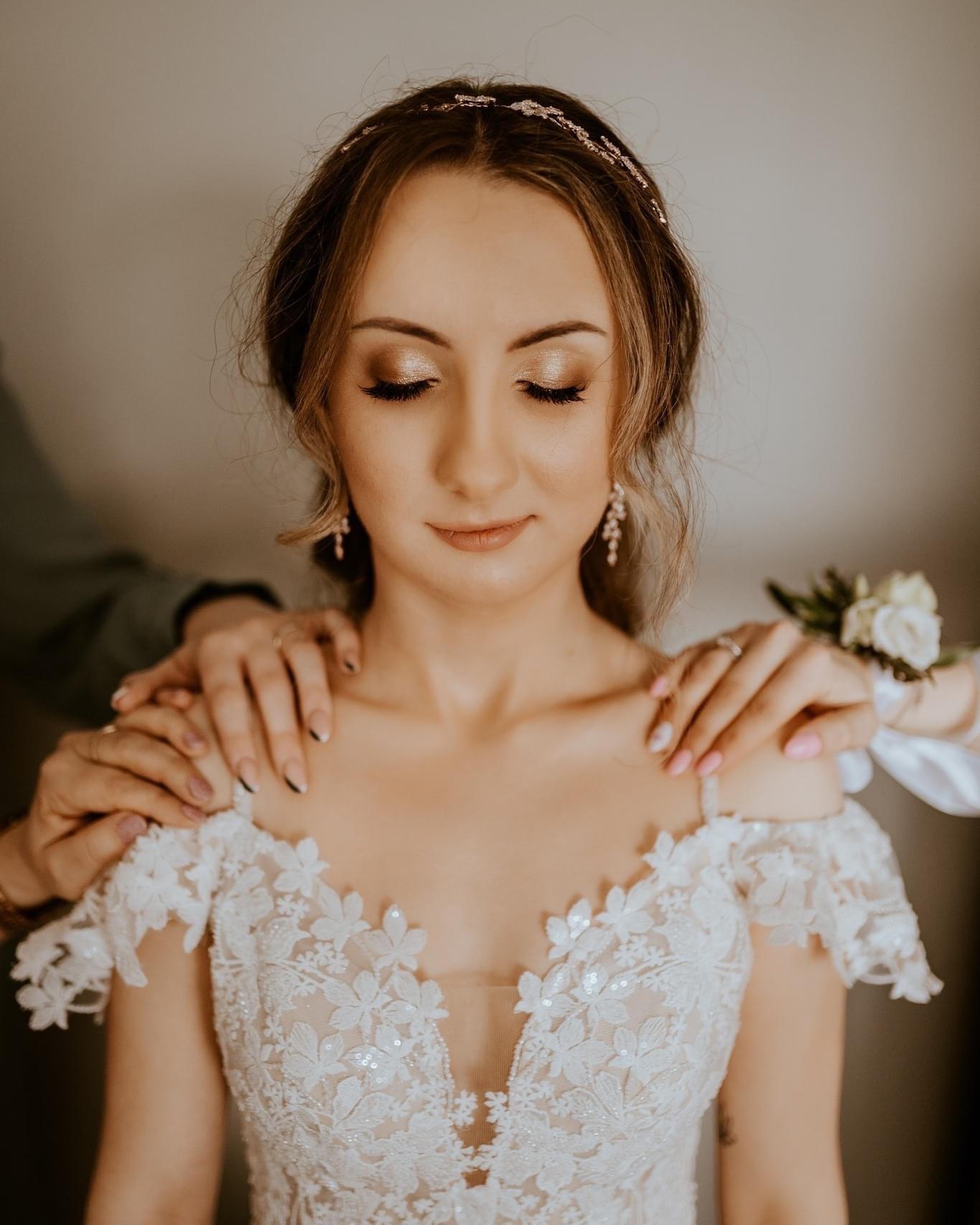 Panna młoda w sukni z koronkowymi aplikacjami, z zamkniętymi oczami, przygotowywana do ślubu przez dwie osoby. Delikatny makijaż i biżuteria.