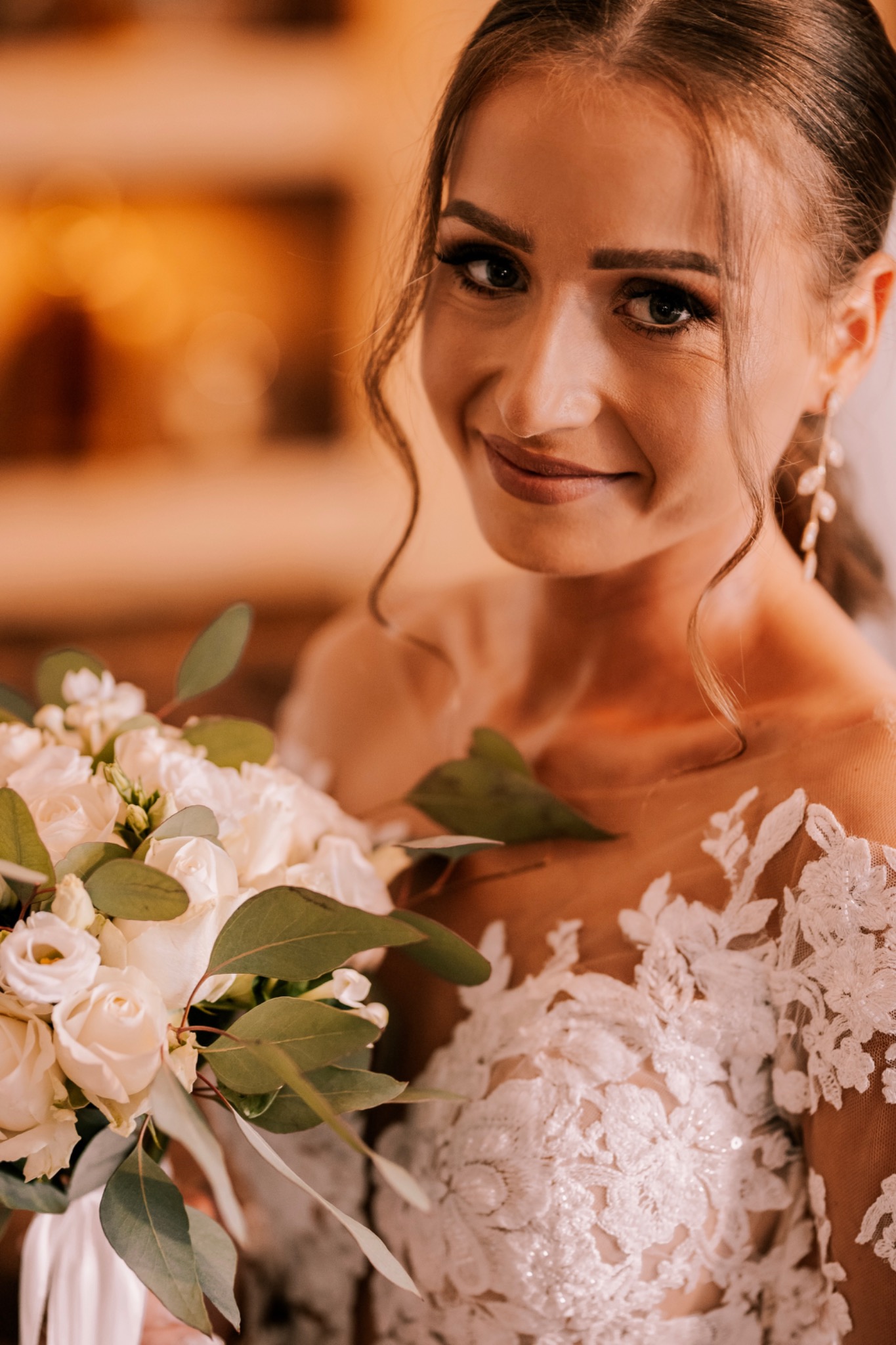 Uśmiechnięta panna młoda w koronkowej sukni trzyma bukiet białych róż i eukaliptusa, delikatne kolczyki wiszące, ciepłe oświetlenie.
