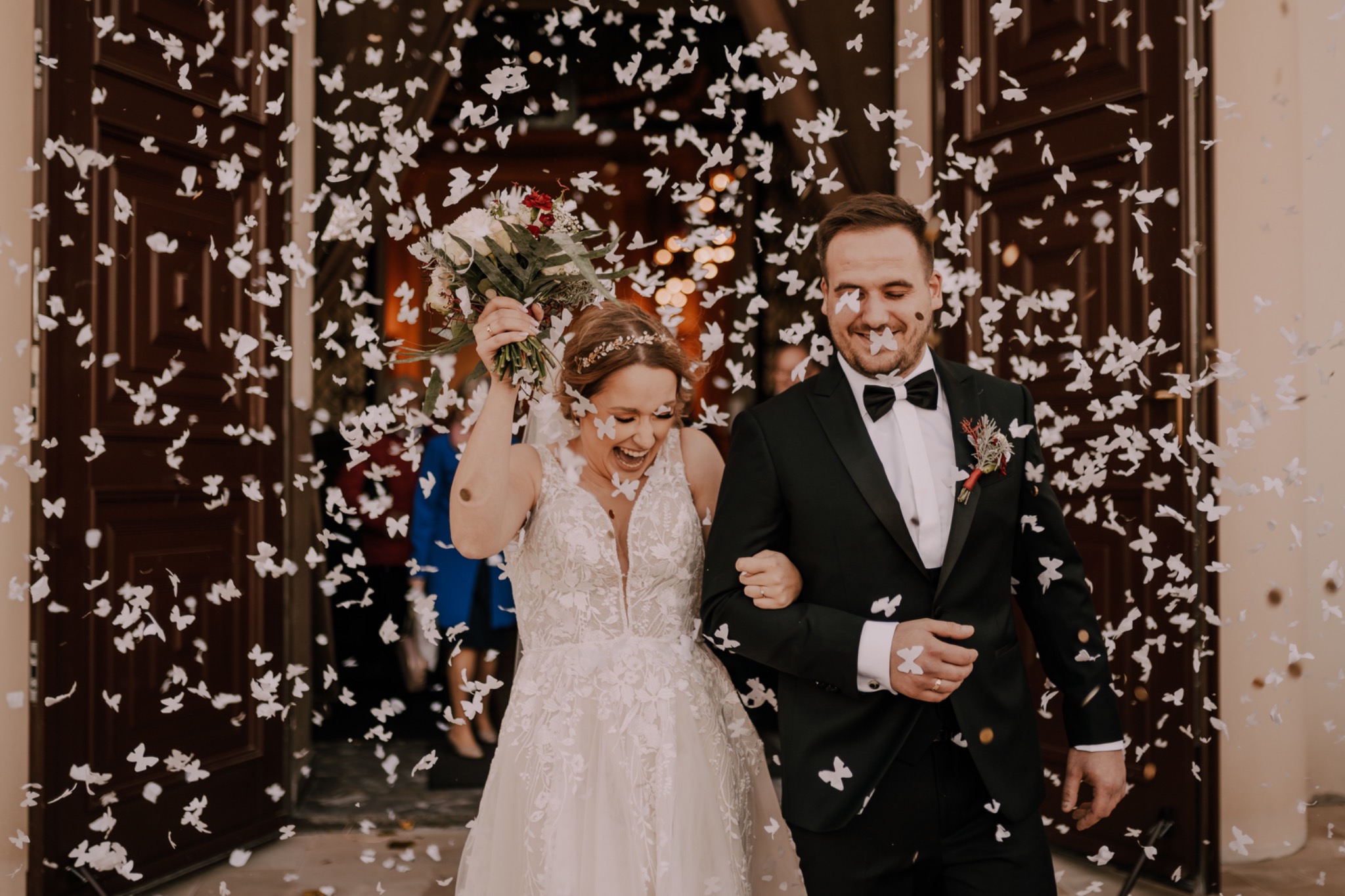 Uśmiechnięta para młoda opuszcza kościół pod gradem białych konfetti w kształcie motyli, panna młoda w koronkowej sukni trzyma bukiet, pan młody w czarnym smokingu z butonierką.