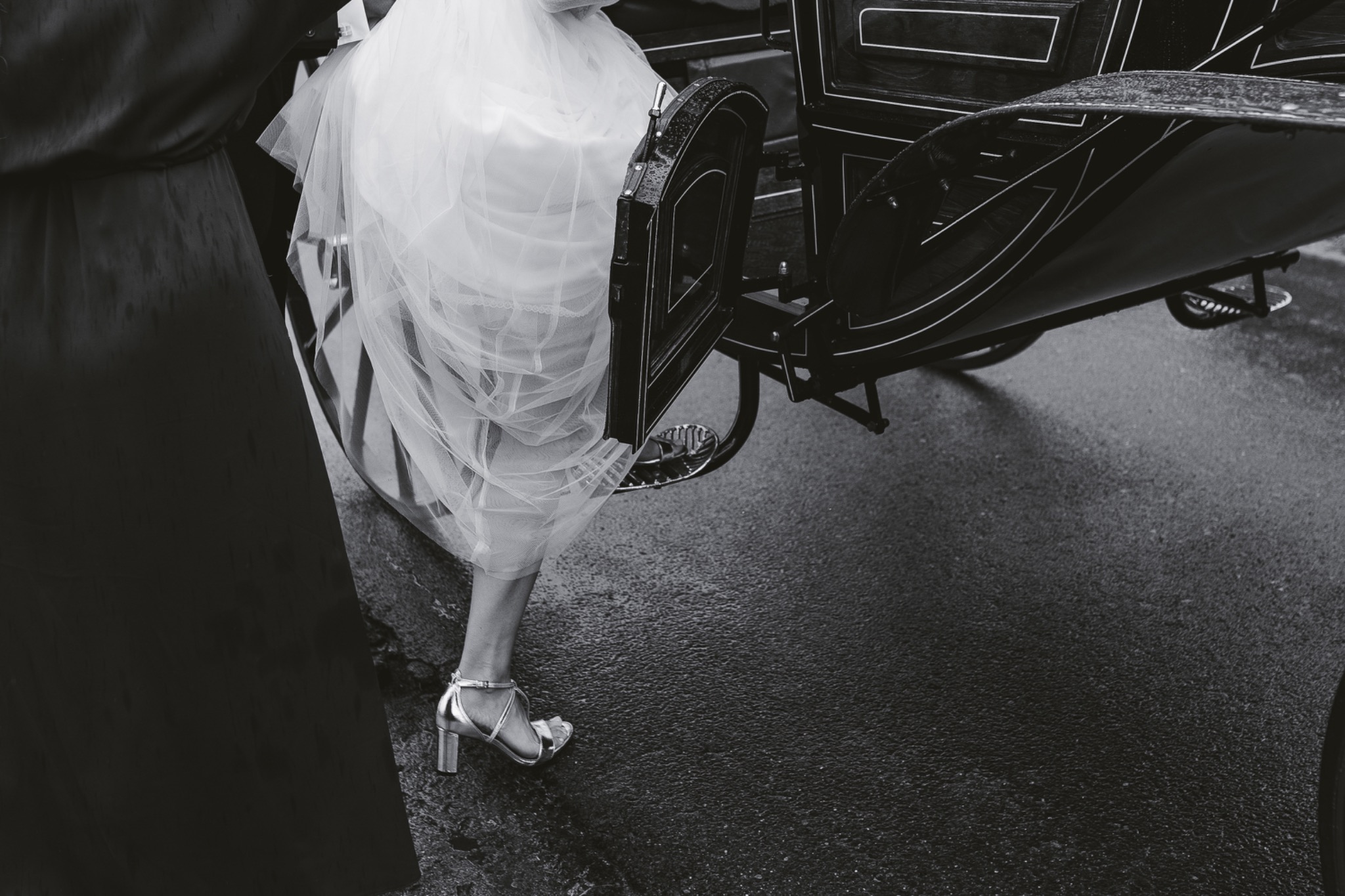 Panna młoda w srebrnych butach wsiada do czarnej karocy w deszczowy dzień, widoczne fragmenty sukni i asystująca osoba.