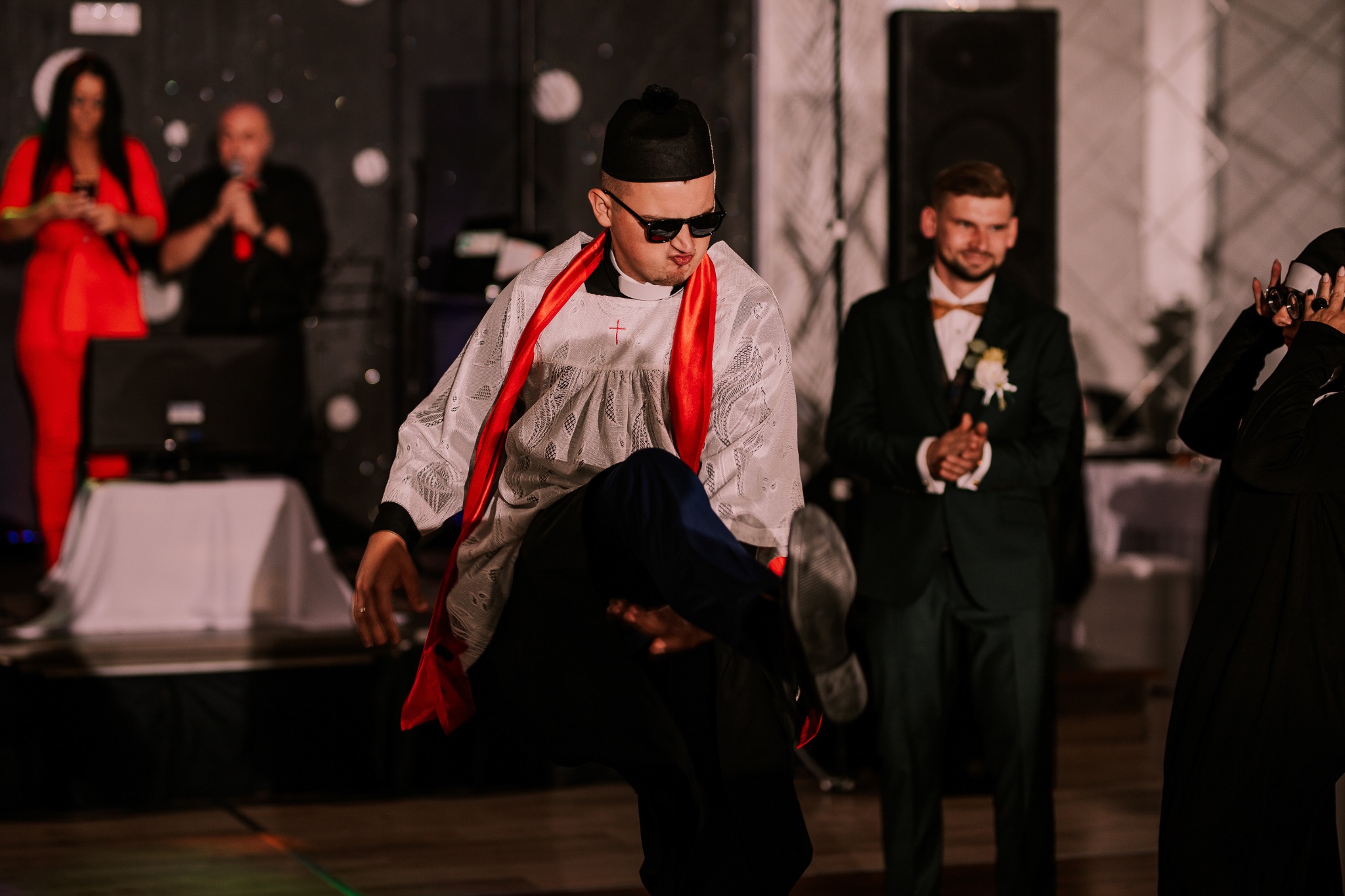 Mężczyzna w przebraniu księdza tańczy na parkiecie podczas imprezy, w tle wodzirej i goście.