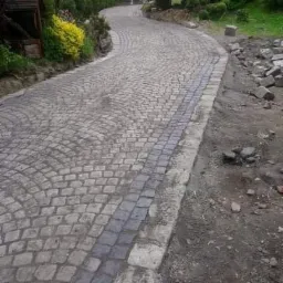 Kręta ścieżka z jasnoszarej kostki granitowej ułożonej w wachlarz, z ciemnoszarym pasem kostki pośrodku, prowadząca przez zielony ogród.