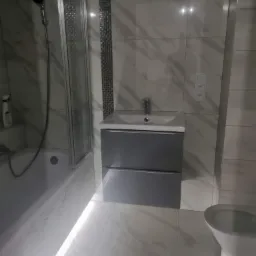 Nowoczesna łazienka z marmurowymi płytkami, szarą szafką pod umywalką, podświetleniem LED przy podłodze i kabiną prysznicową z czarną słuchawką.