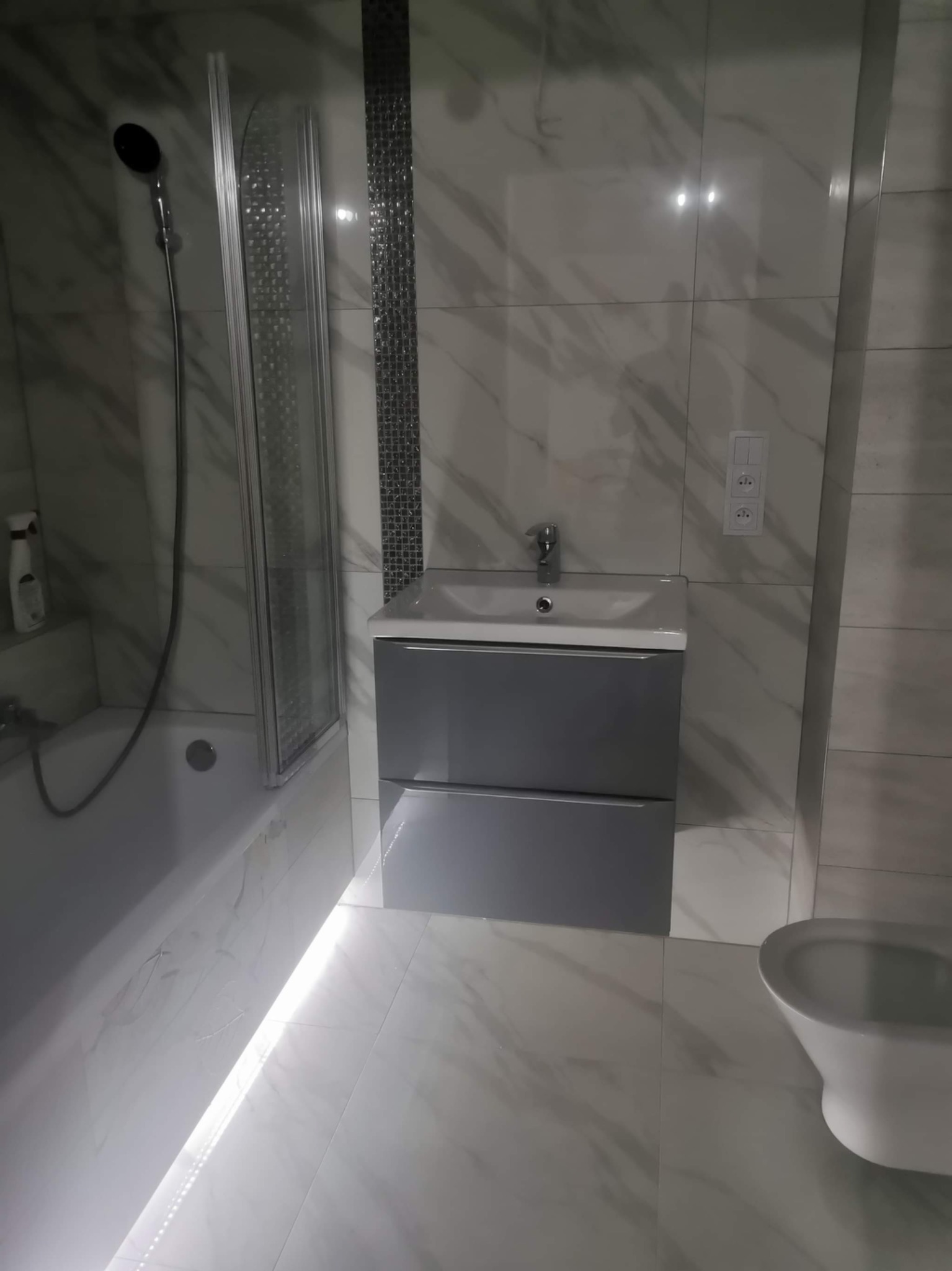 Nowoczesna łazienka z marmurowymi płytkami, szarą szafką pod umywalką, podświetleniem LED przy podłodze i kabiną prysznicową z czarną słuchawką.