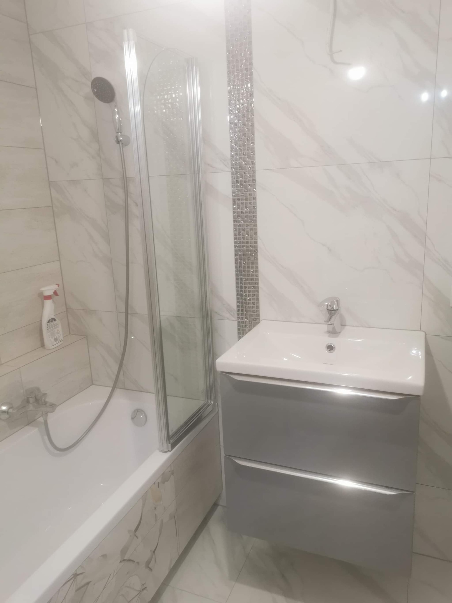 Nowoczesna łazienka po remoncie: białe marmurowe płytki, kabina prysznicowa z pojedynczą ścianką ze szkła, umywalka z szarą szafką z dwoma szufladami, dekoracyjny pas mozaiki na ścianie.