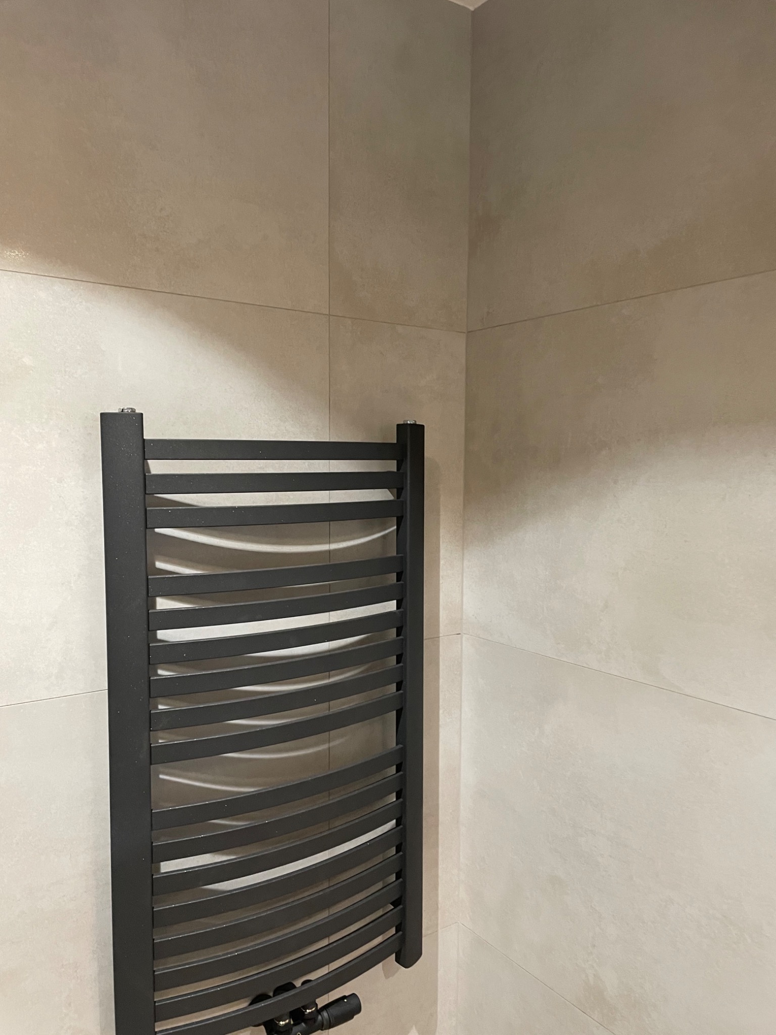 Czarny, drabinkowy grzejnik łazienkowy zamontowany na beżowych, matowych płytkach w rogu pomieszczenia.