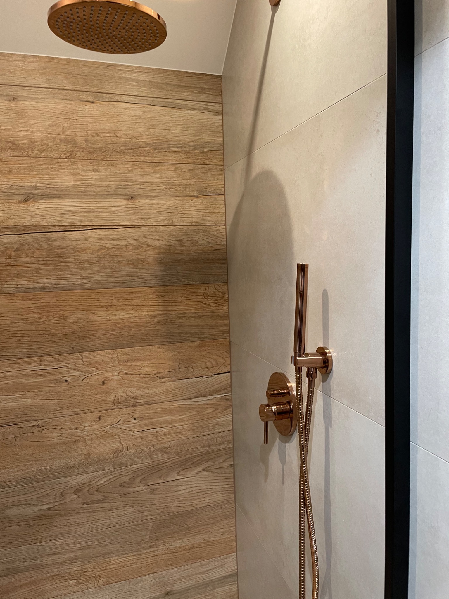 Nowoczesna kabina prysznicowa z deszczownicą i baterią w kolorze różowego złota, wykończona płytkami imitującymi drewno i jasnymi płytkami ceramicznymi.