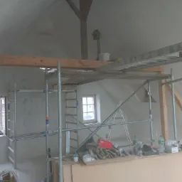 Poddasze w trakcie prac wykończeniowych: widoczne rusztowanie, drabiny, narzędzia i materiały budowlane, ściany przygotowane do malowania, okna z widokiem na czerwoną elewację budynku.