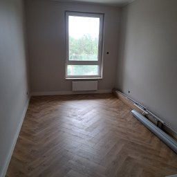 ROB-BUD Prestige wykończenia wnętrz Robert Makowski - Pusty pokój z nową podłogą ułożoną we wzór jodełki, grzejnikiem pod oknem i listwami przypodłogowymi w opakowaniu przy ścianie.