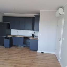 ROB-BUD Prestige wykończenia wnętrz Robert Makowski - Niebieska zabudowa kuchenna z białymi płytkami metra, drewnianym blatem i częściowo zamontowanymi szafkami, widok na podłogę z paneli i białe drzwi w nowym mieszkaniu.