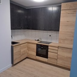 ROB-BUD Prestige wykończenia wnętrz Robert Makowski - Częściowo zrealizowana zabudowa kuchenna z drewnianymi frontami szafek dolnych i czarnymi frontami szafek górnych, białe płytki na ścianie, czarny zlew i płyta indukcyjna.