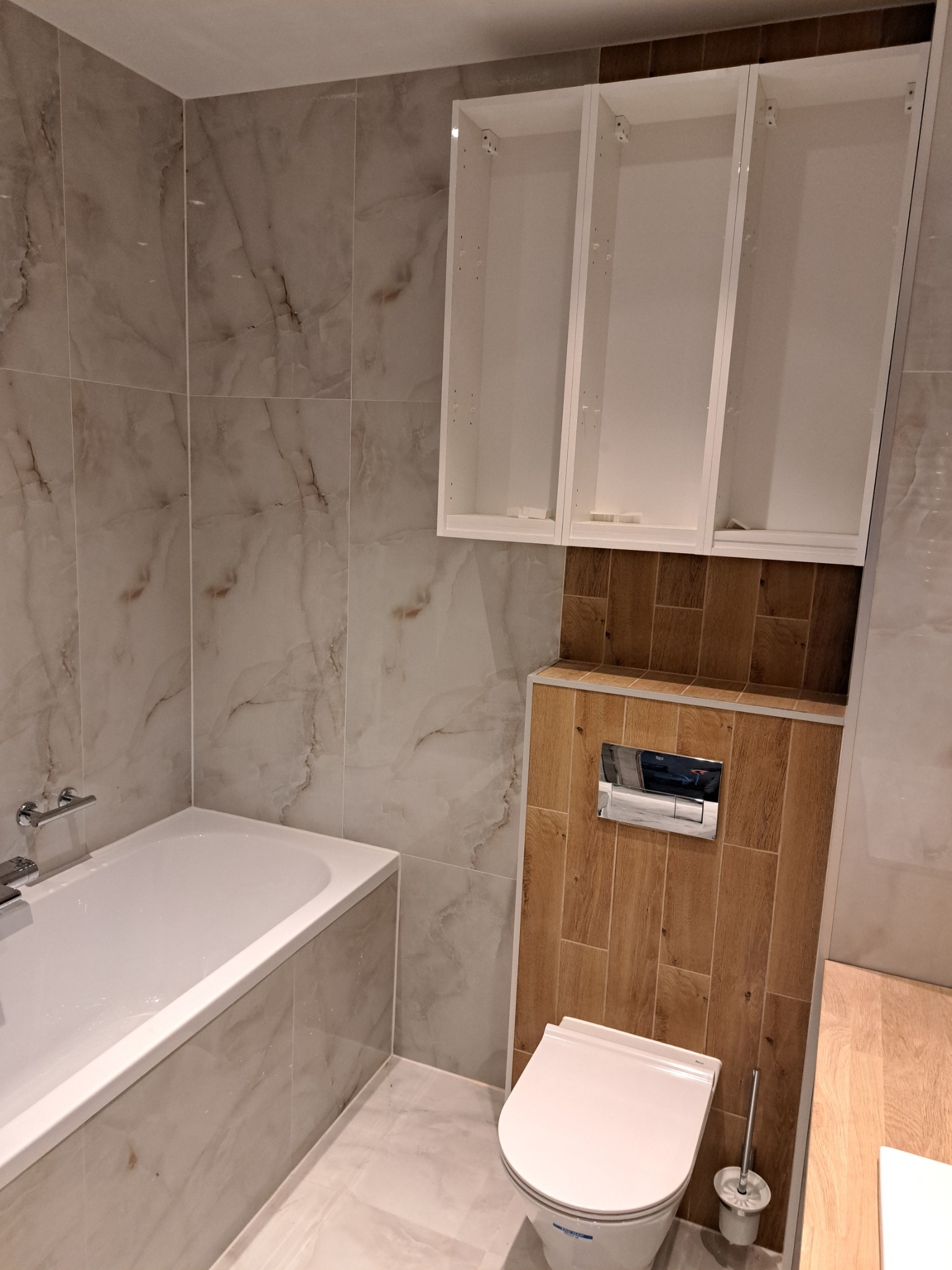 Wykończona łazienka z marmurowymi płytkami, wiszącą toaletą, drewnopodobnymi panelami, białymi szafkami bez frontów i wanną z baterią.