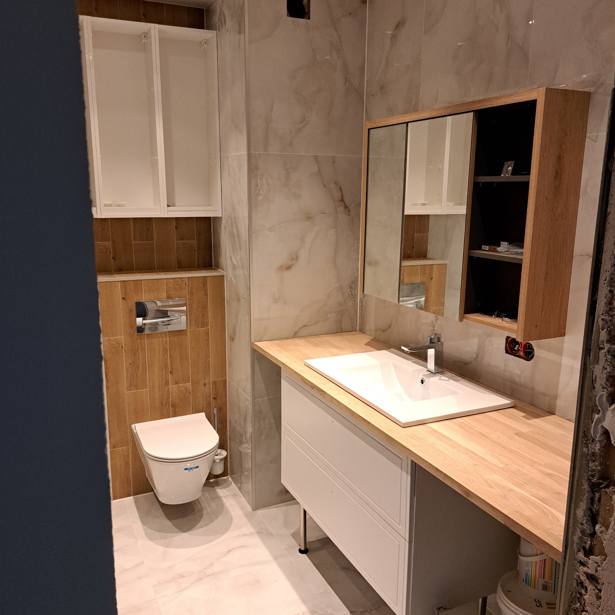 Wnętrze łazienki w trakcie prac wykończeniowych, widoczna podwieszana toaleta, umywalka z szafką pod blatem z jasnego drewna, lustro w drewnianej ramie z otwartymi półkami, marmurowe płytki...