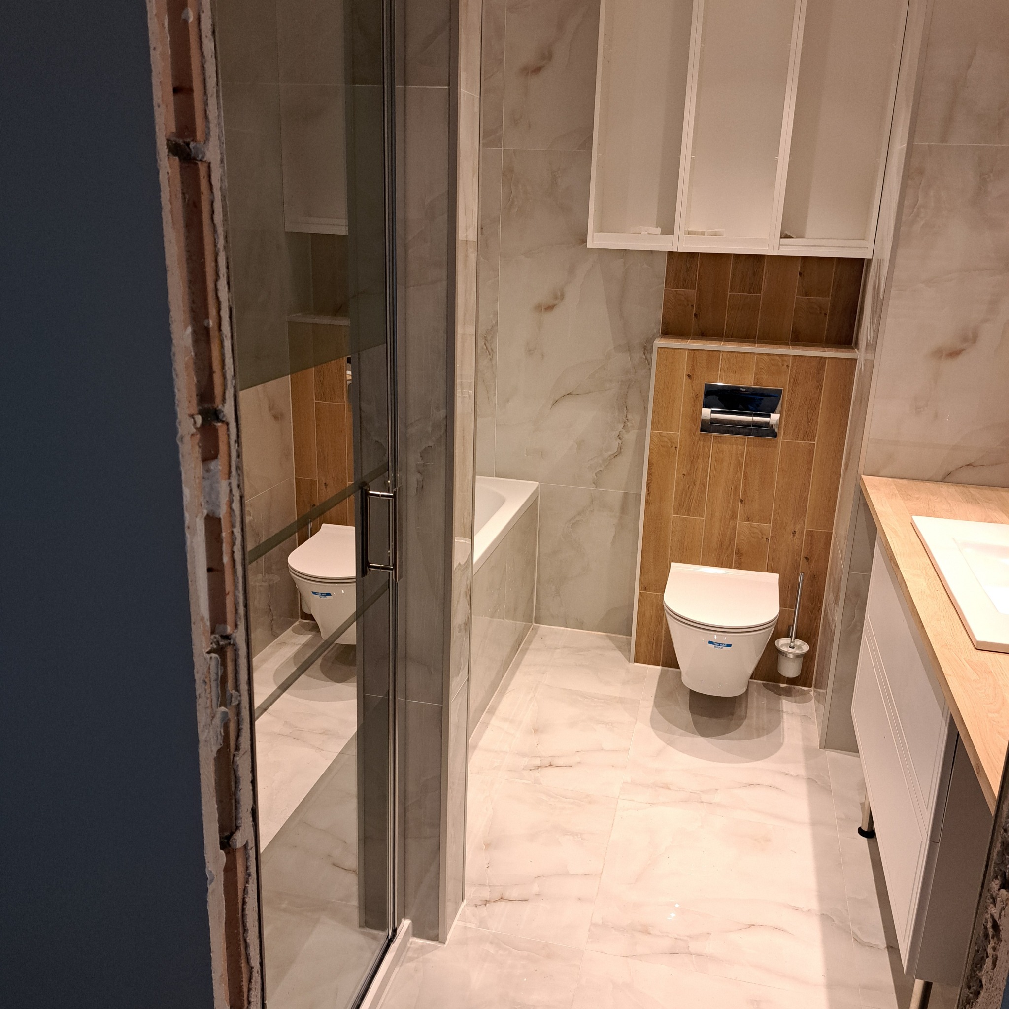 Wykończona łazienka z marmurowymi płytkami, kabiną prysznicową z ciemnymi ramami, wiszącą toaletą i szafkami w kolorze drewna, widoczna surowa ściana w trakcie prac wykończeniowych.