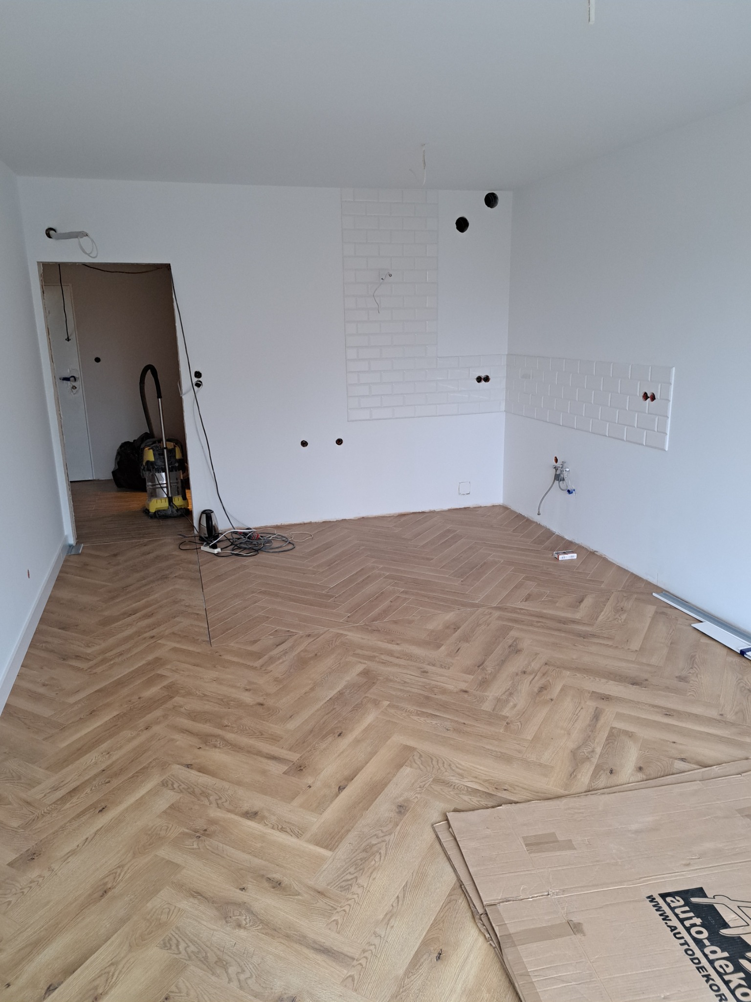 Kuchnia w trakcie remontu: ułożona podłoga z paneli w jodełkę, częściowo ułożone białe płytki metro na ścianie, widoczne instalacje, odkurzacz i kartony na podłodze.