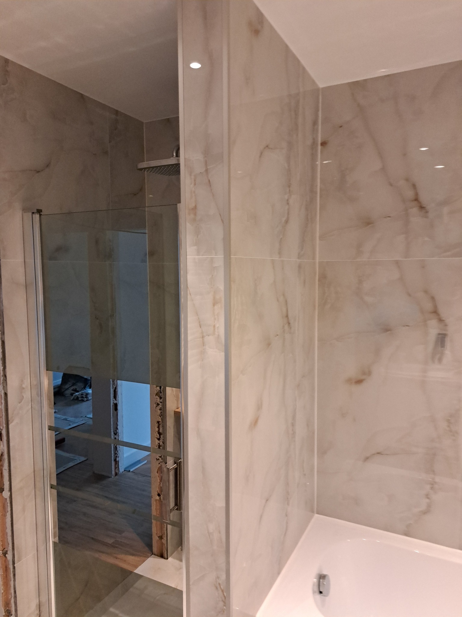 Wykończona łazienka z beżowymi płytkami imitującymi marmur, kabina prysznicowa ze srebrnym uchwytem, widoczny fragment wanny.
