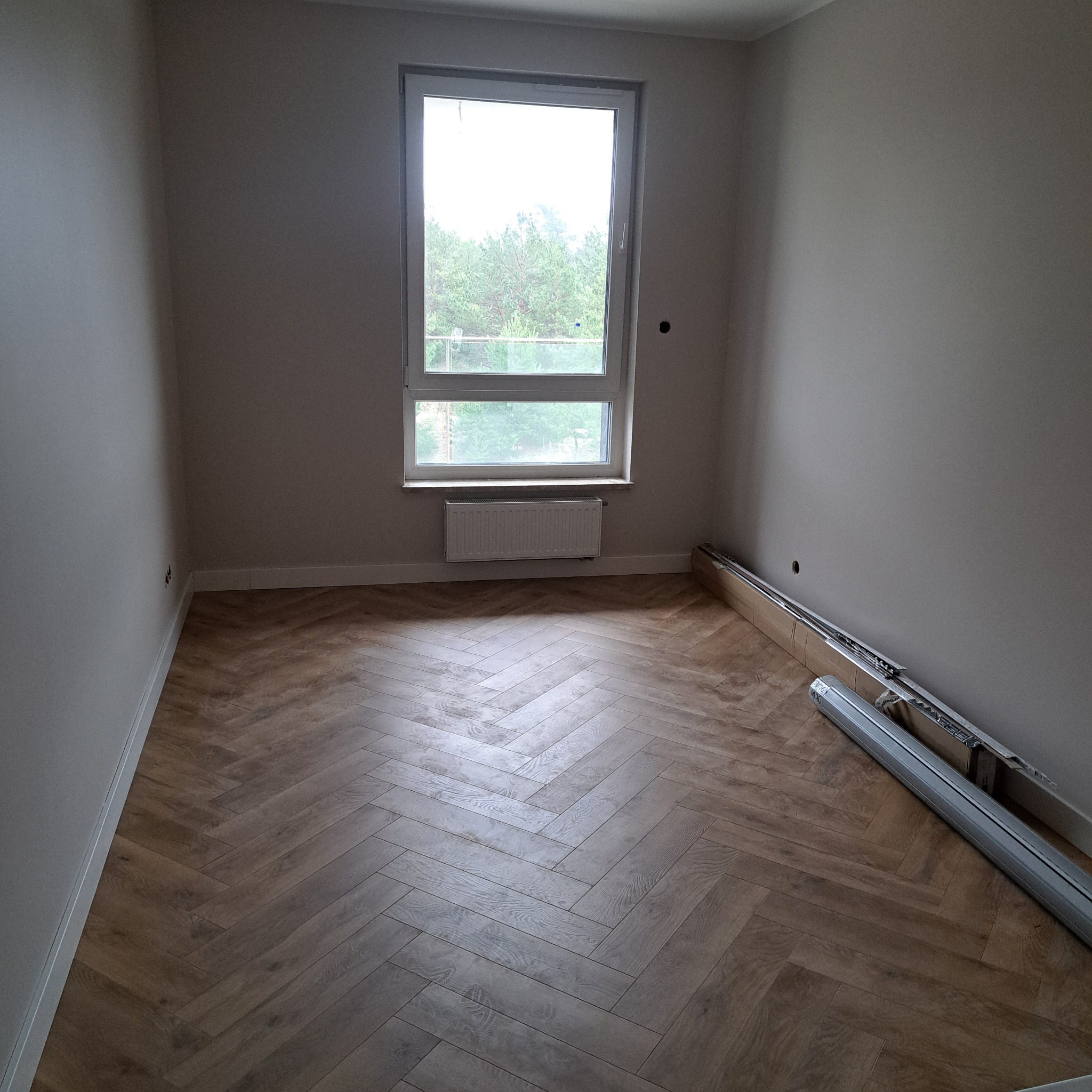 Pusty pokój z nową podłogą ułożoną we wzór jodełki, grzejnikiem pod oknem i listwami przypodłogowymi w opakowaniu przy ścianie.