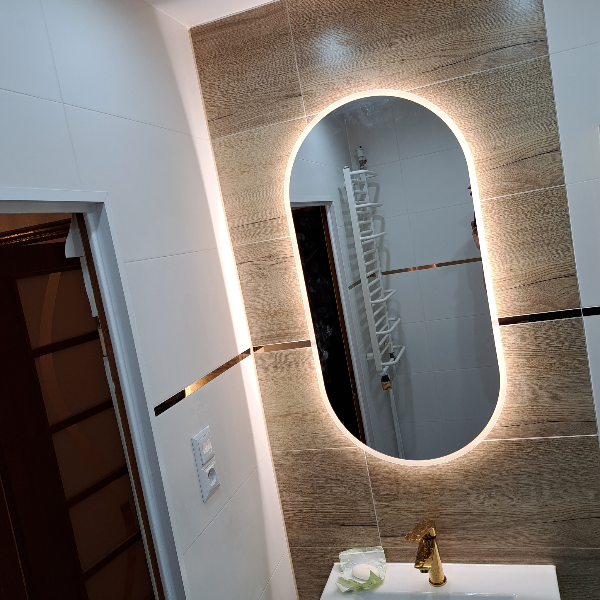 Nowoczesna łazienka z owalnym lustrem podświetlanym LED, drewnopodobnymi płytkami i złotą armaturą, widok na grzejnik drabinkowy w odbiciu lustra.