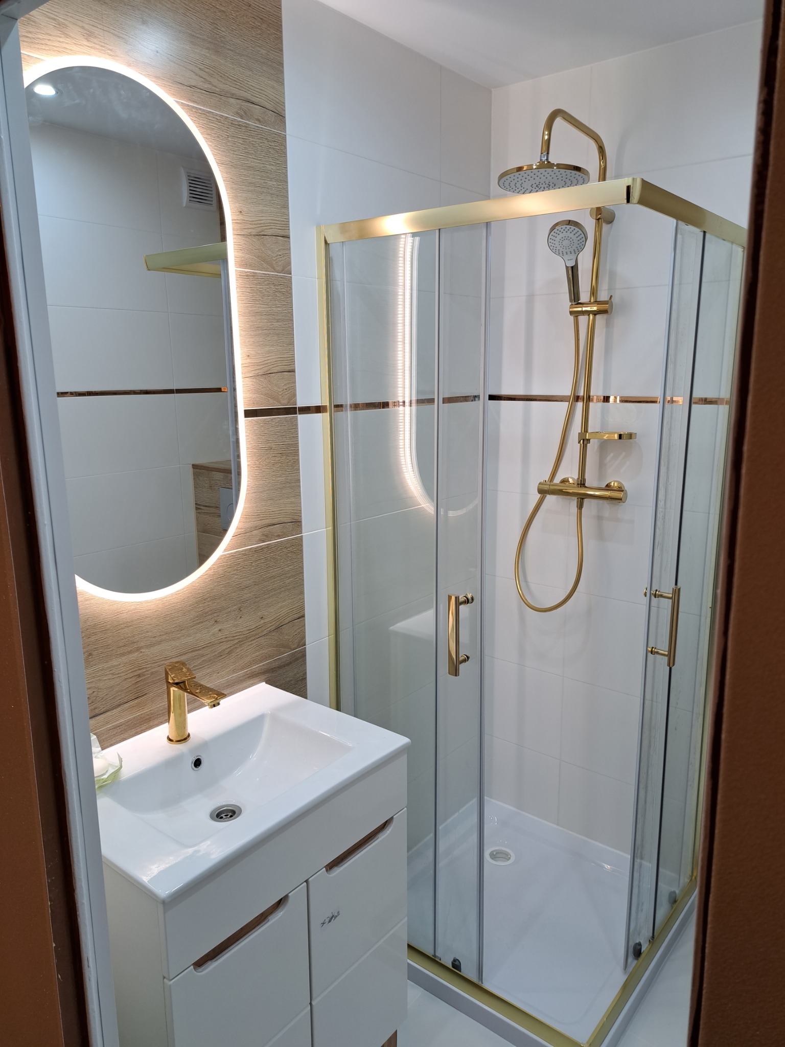 Nowoczesna łazienka z kabiną prysznicową i umywalką, złote akcenty na baterii, ramie kabiny i armaturze, owalne lustro z podświetleniem LED, białe płytki imitujące drewno.